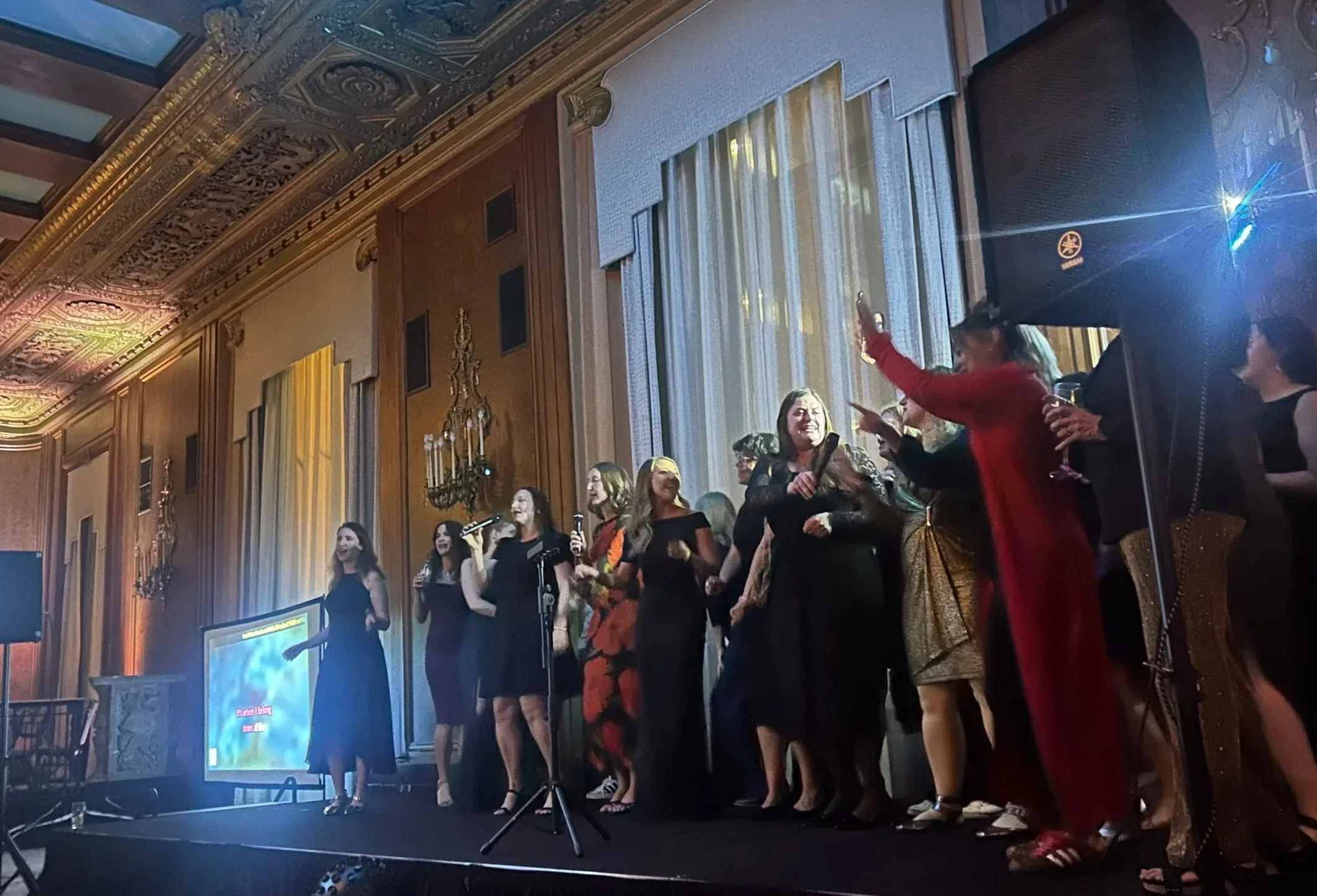 A group of women singing on stage at an elegant venue with ornate wood paneling, chandeliers, and large windows, during a performance or karaoke event.