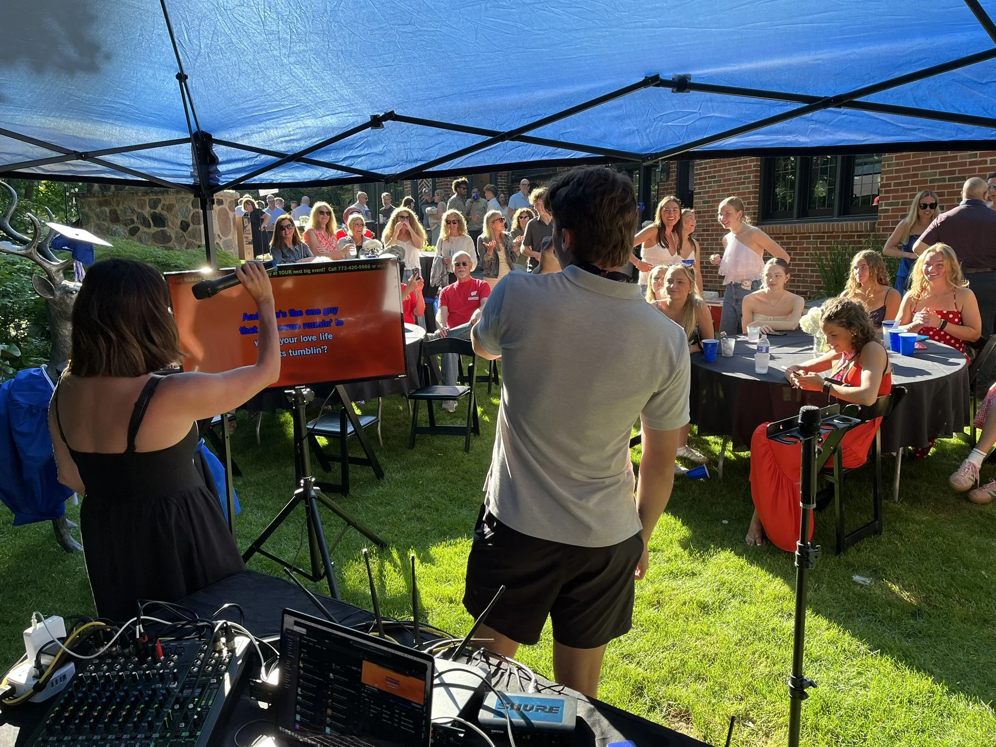 A group of people gathered outdoors under a blue canopy, some seated around tables with drinks, others standing and socializing, with a person in the foreground operating audio equipment.
