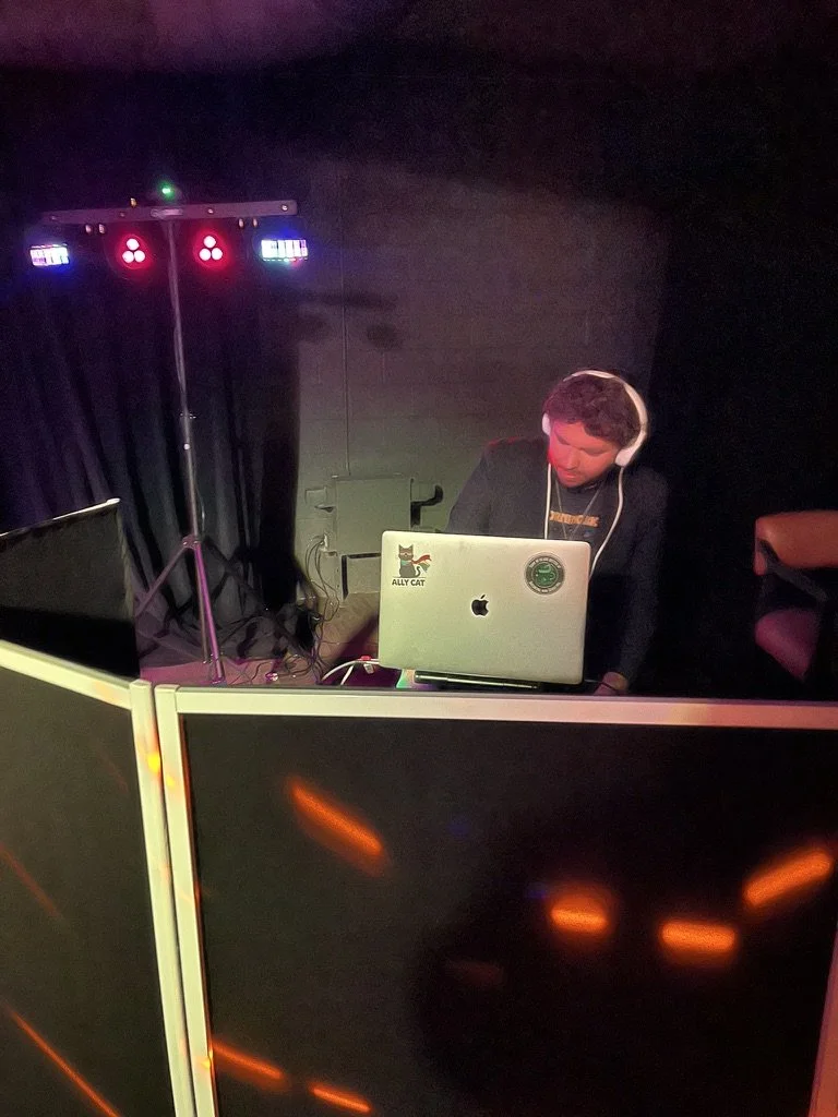 Person with headphones operating DJ equipment, including a laptop, in a dark setting with colorful lights.
