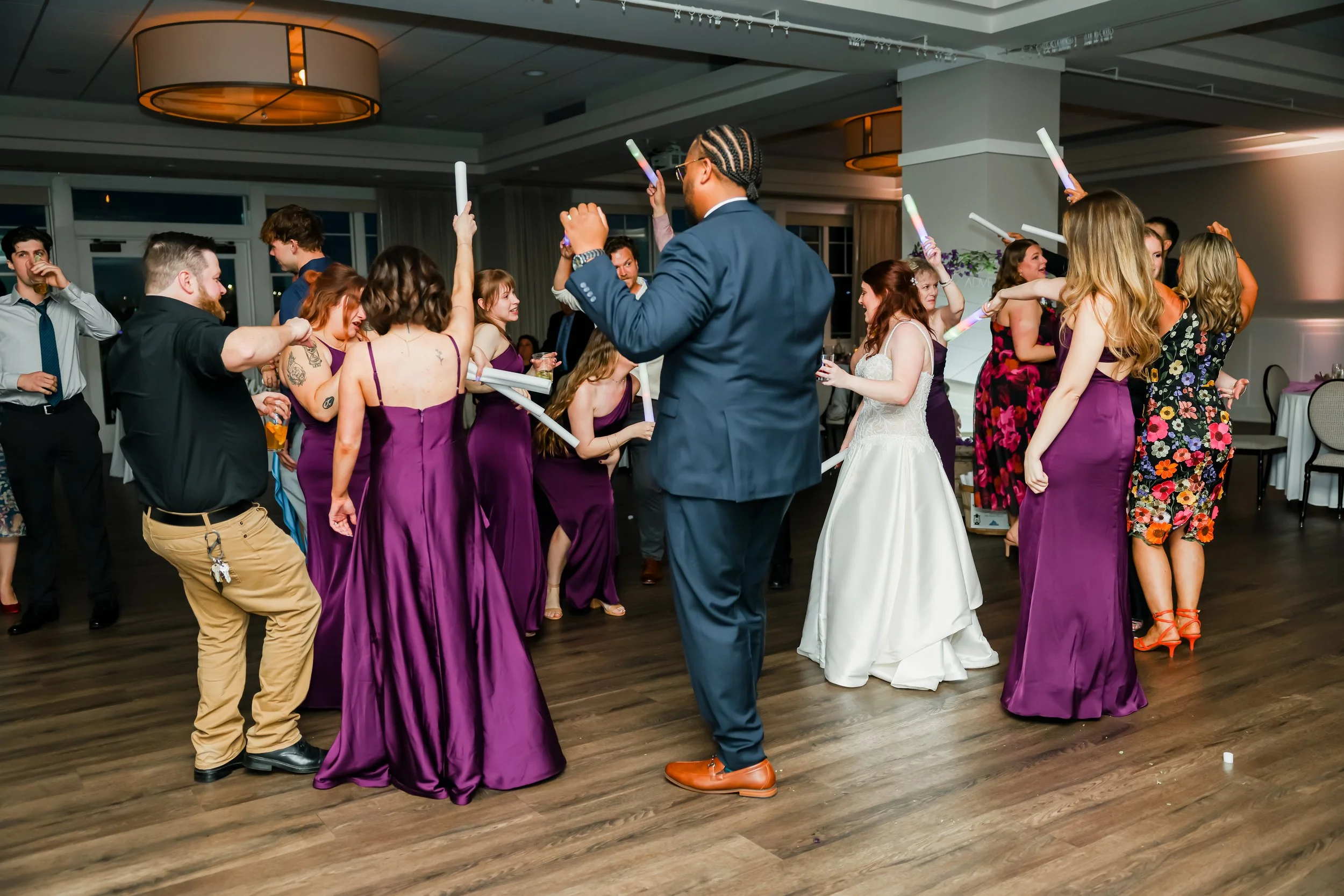 People dancing at a wedding reception, with women in purple dresses and a bride in a white dress, while a man in a suit and tie leads them in a dance.