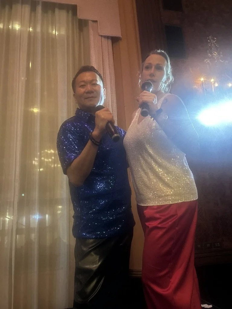 Two people standing on a stage holding microphones, dressed in shiny and colorful clothing, near a window with curtains and warm lighting.
