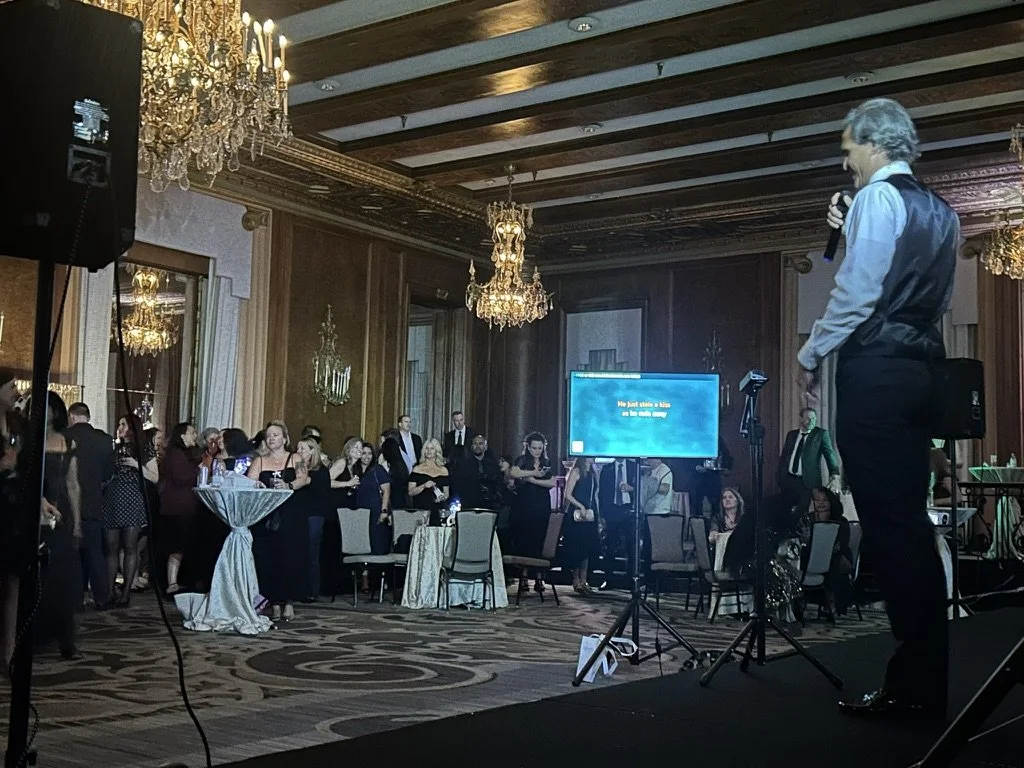 A man in formal attire is speaking into a microphone on a stage at a banquet or event hall, with an audience of people standing and sitting at tables, some dancing and socializing, in a room with chandeliers and wood-paneled walls.