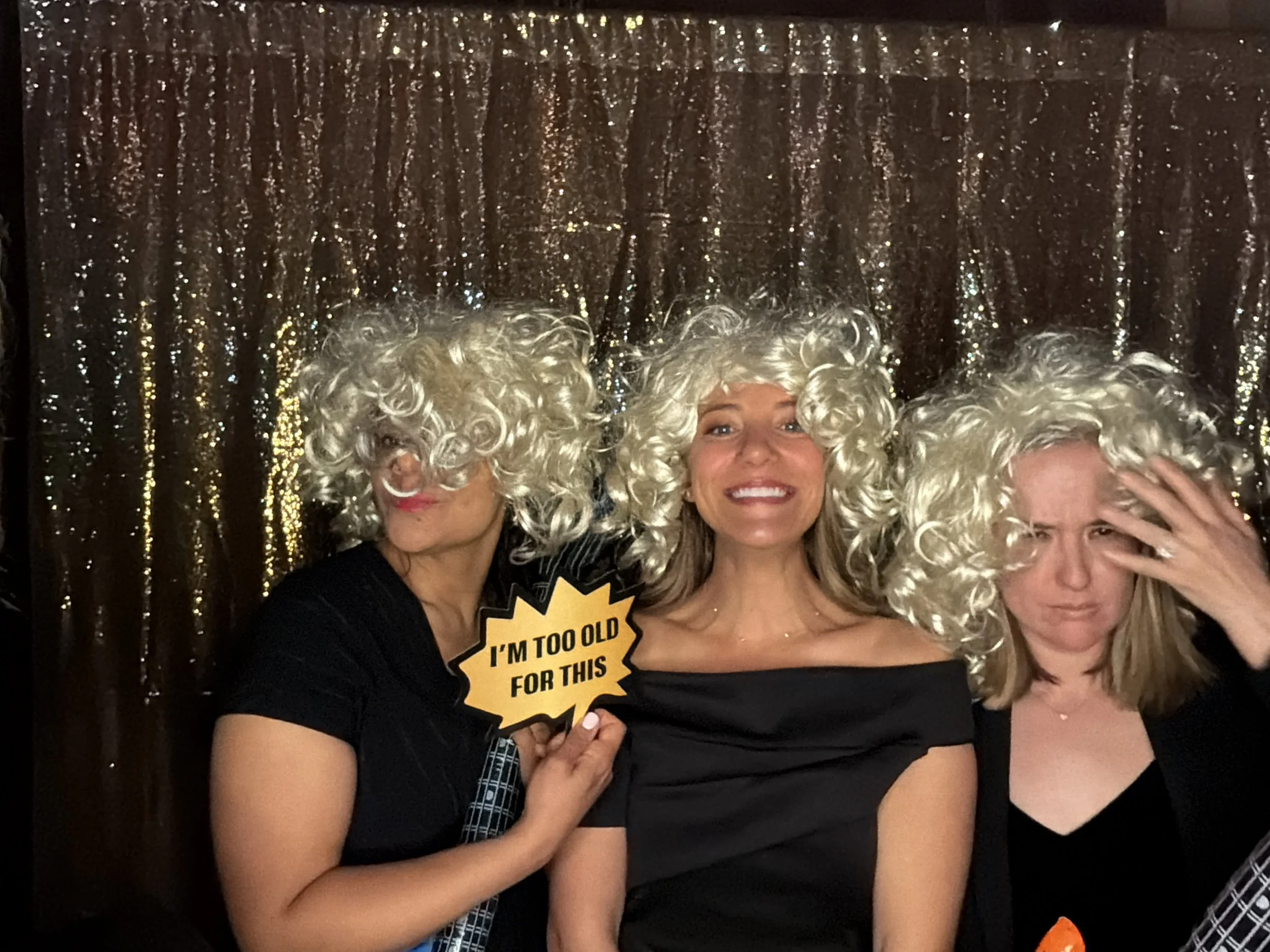Three women in curly white wigs smiling at a photo booth. The woman in the center is holding a sign that reads 'I'M TOO OLD FOR THIS.' The women are wearing black outfits, and the background is a sparkly gold curtain.