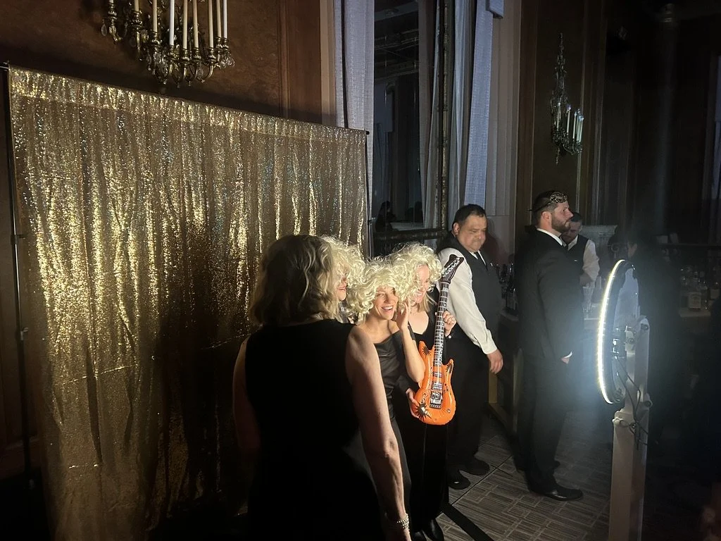 A group of people dressed in formal attire at a party or event, standing near a gold-colored curtain with a chandelier above. One woman is holding a small orange electric guitar prop, and they are positioned in front of a photo booth with a bright ri