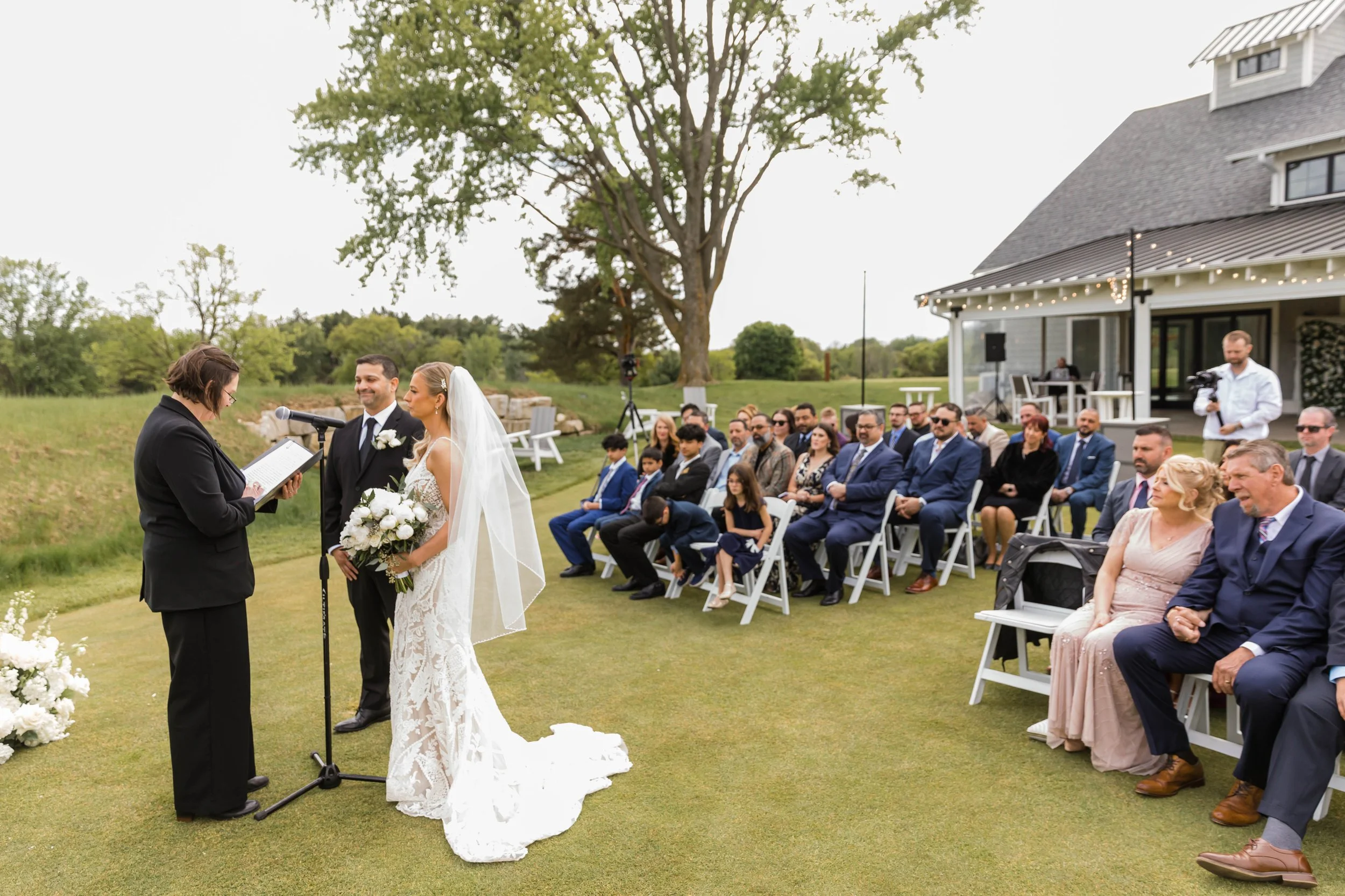 Outdoor wedding ceremony with a bride and groom standing before an officiant, surrounded by seated guests on a grassy lawn with trees and a house in the background.