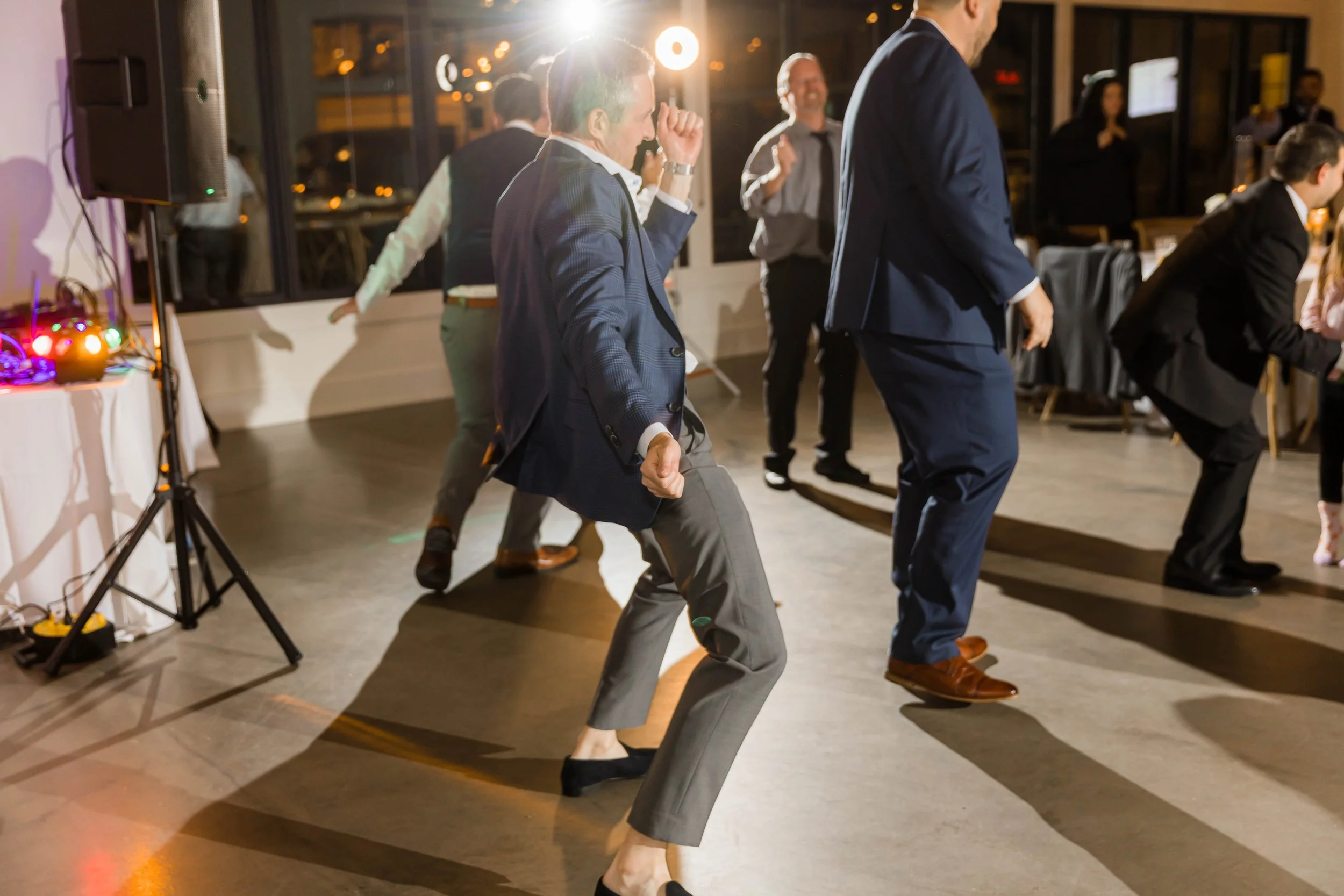 People dancing at an indoor event, dressed in formal attire, with some men in suits and women in dresses, enjoying music and socializing.