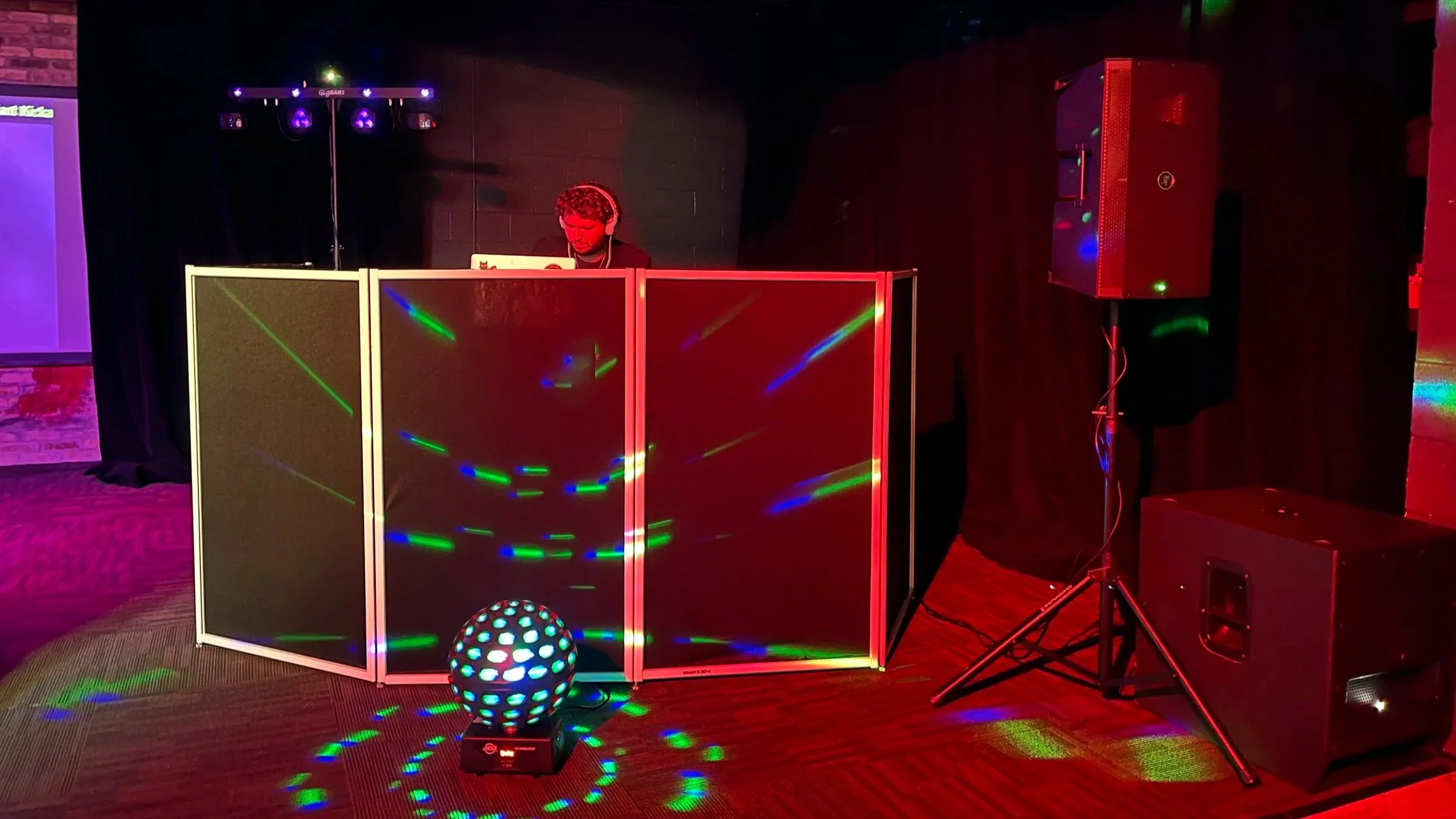 A DJ performing with colorful lights and a disco ball in a dark room, surrounded by speakers and equipment.