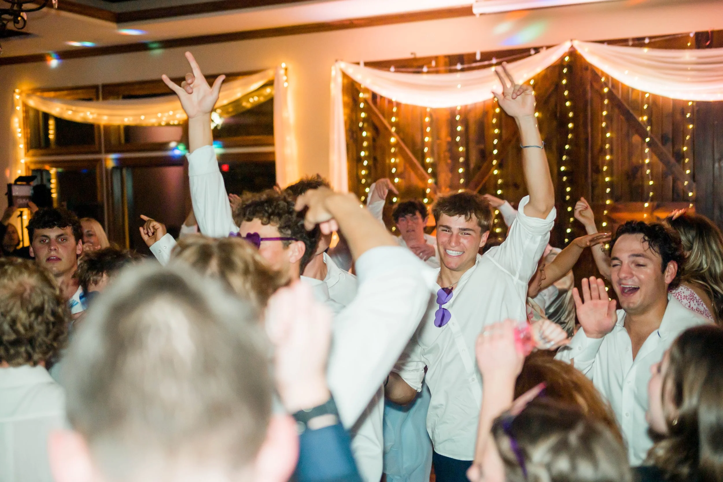 People dancing and celebrating at an indoor party with warm lighting, fairy lights, and draped fabric decorations.