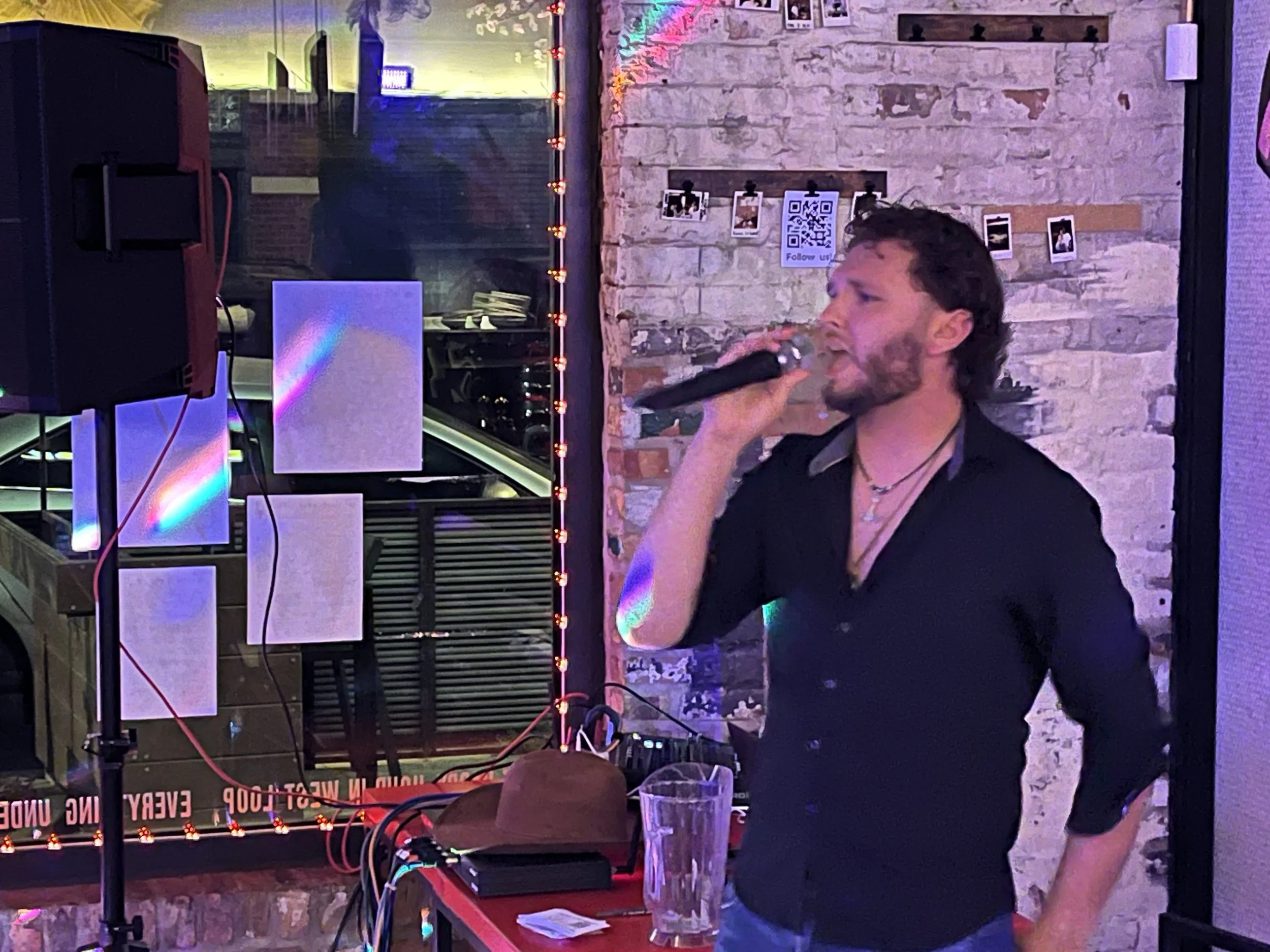 A man singing into a microphone at a party or bar, with a brick wall and colorful lights in the background, next to a DJ setup and a glass of drink on the table.