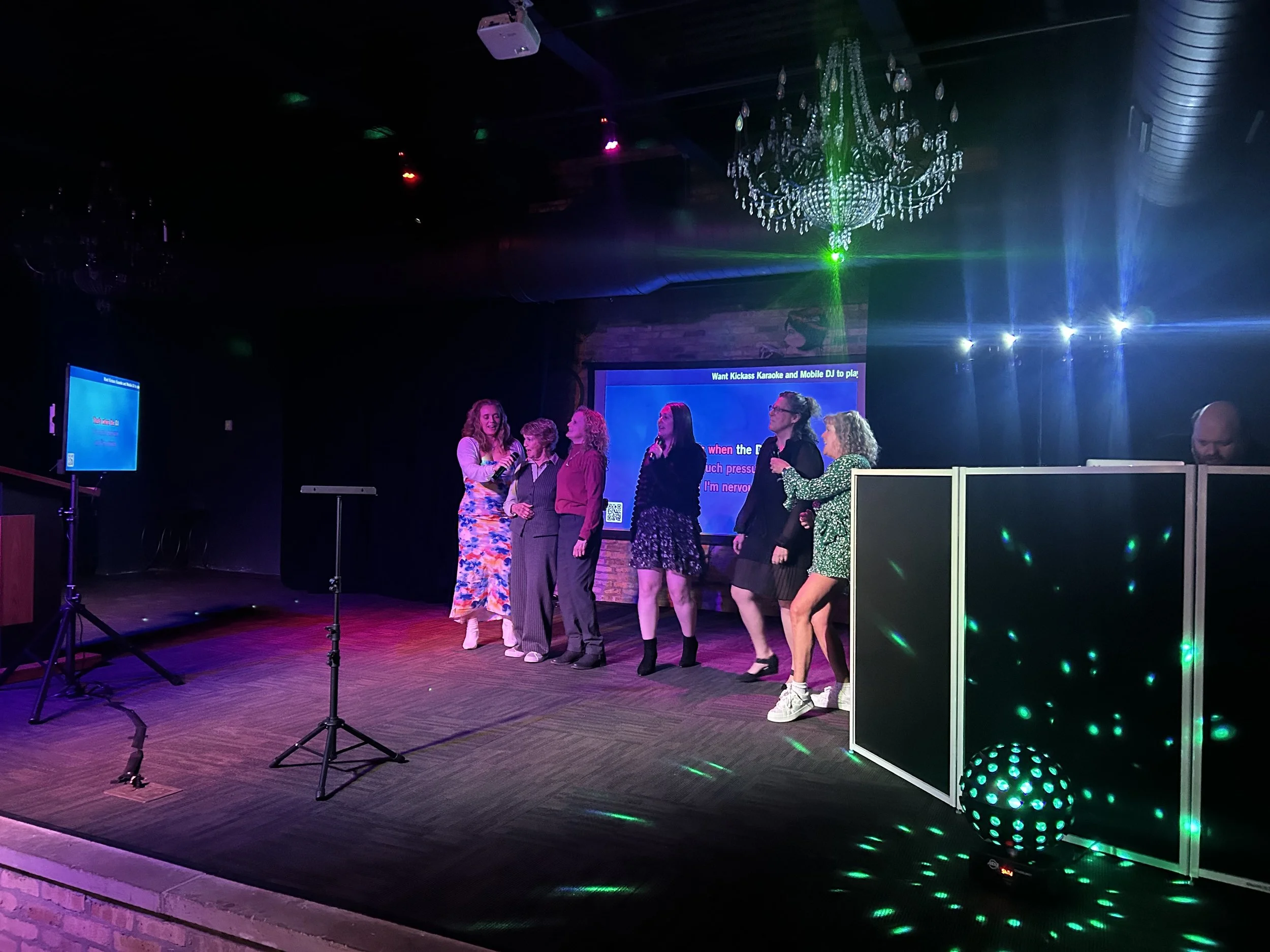 A group of women singing on a stage with a large screen in the background, decorated with a chandelier and colorful lights.