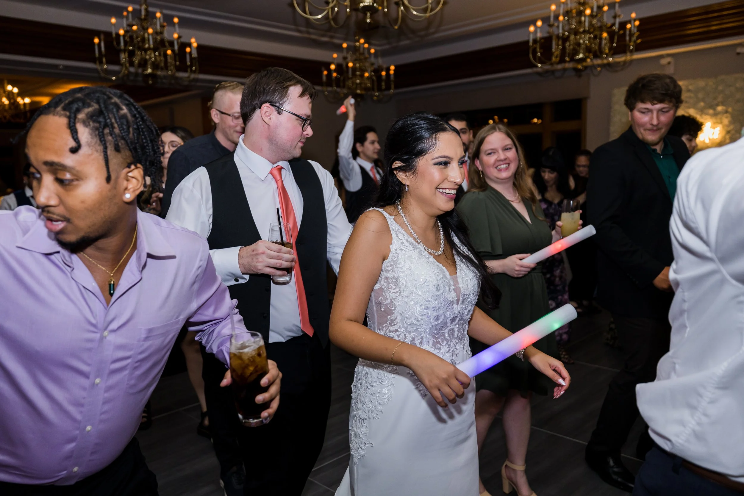 Group of people dancing at a wedding reception, including a woman in a white lace wedding gown, smiling and holding a lit LED light stick, with others around her holding drinks and enjoying the event.