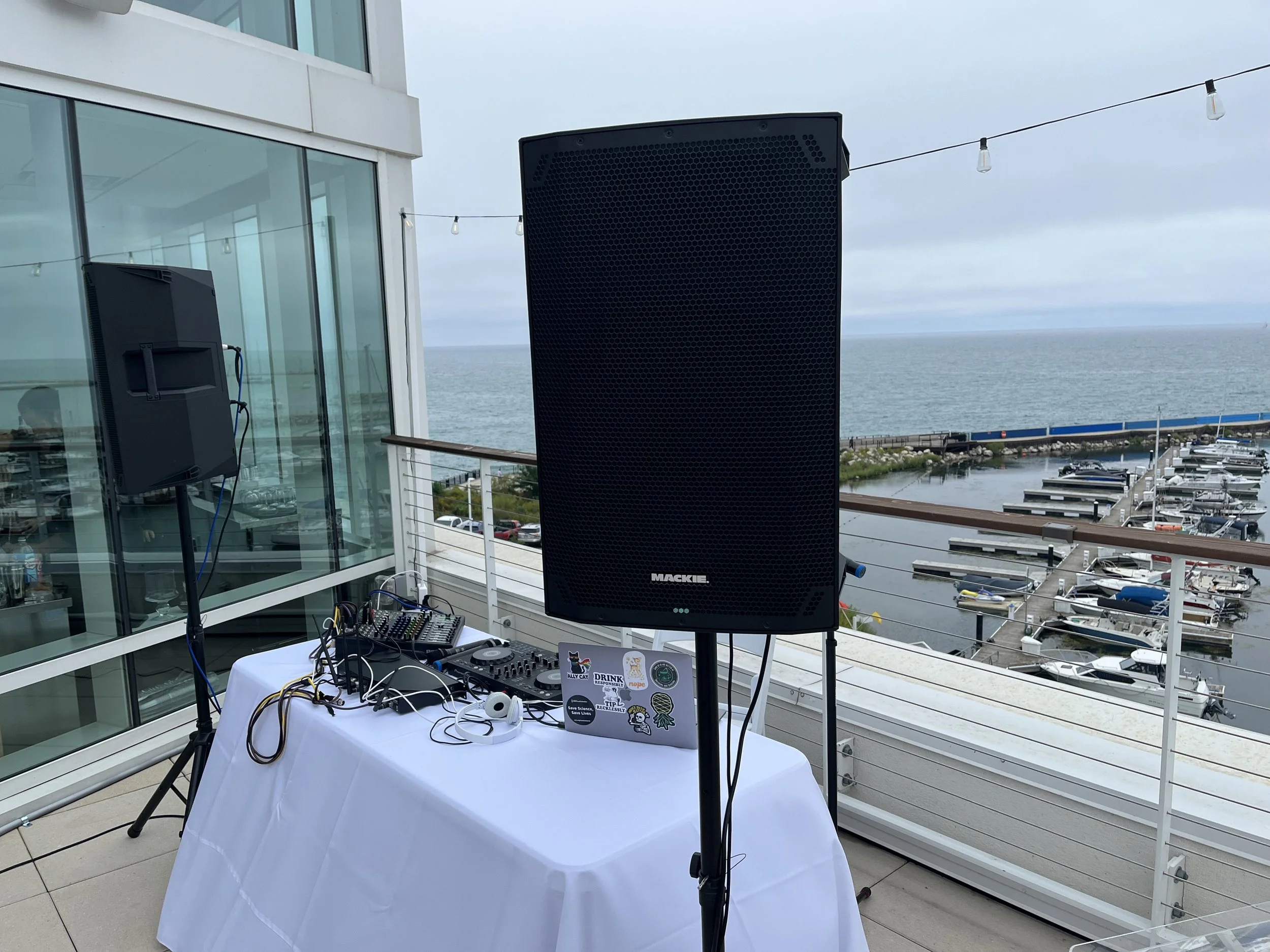 Outdoor event setup with speaker, DJ equipment, and decorative string lights overlooking a marina and ocean.