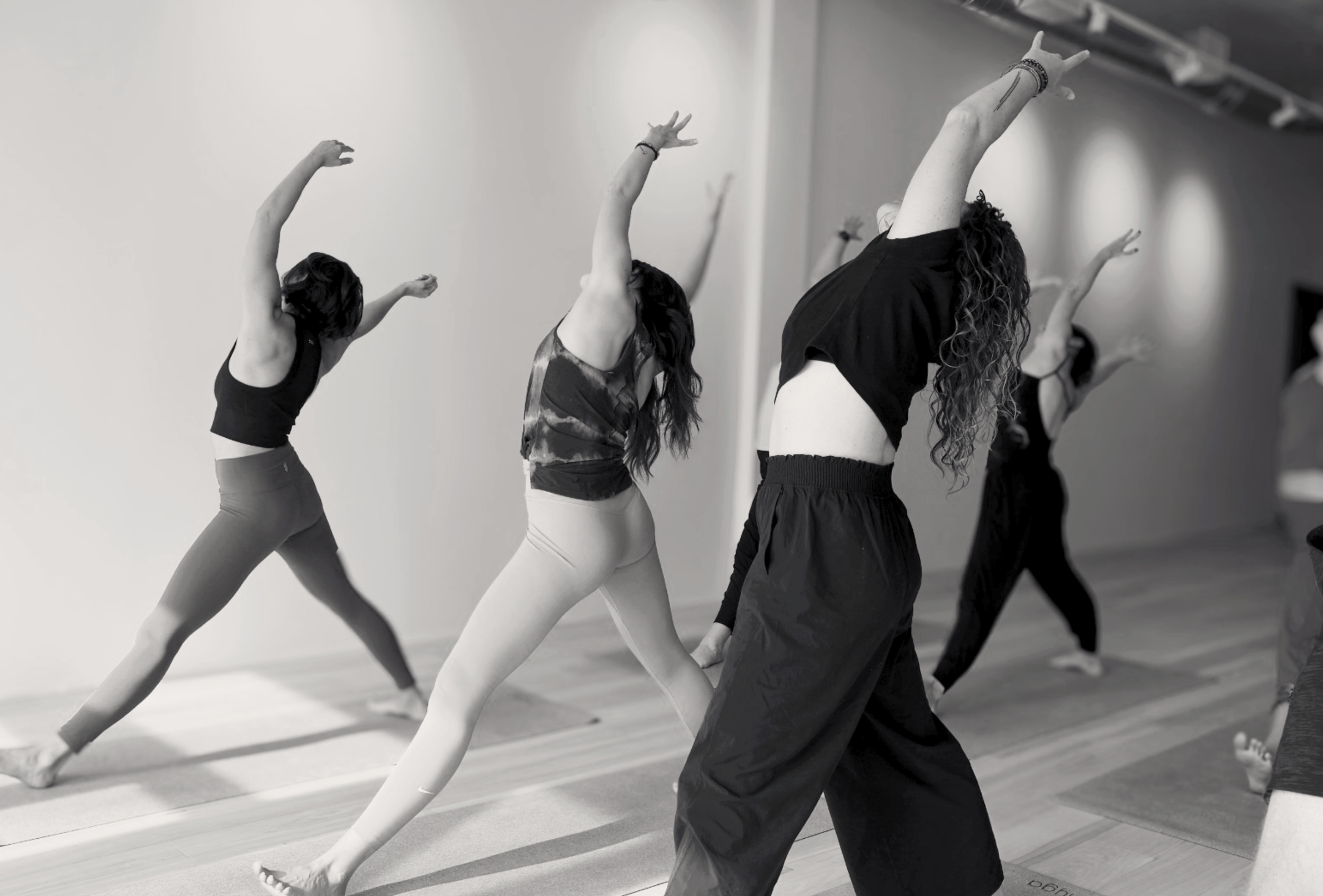 Women in workout clothes stretching in a yoga class, standing in a line with arms raised and leaning to the side.