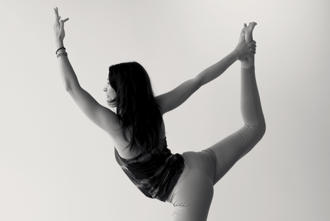 A woman practicing yoga in a pose, standing on one leg and holding her foot behind her back with one hand, extending the other arm forward.