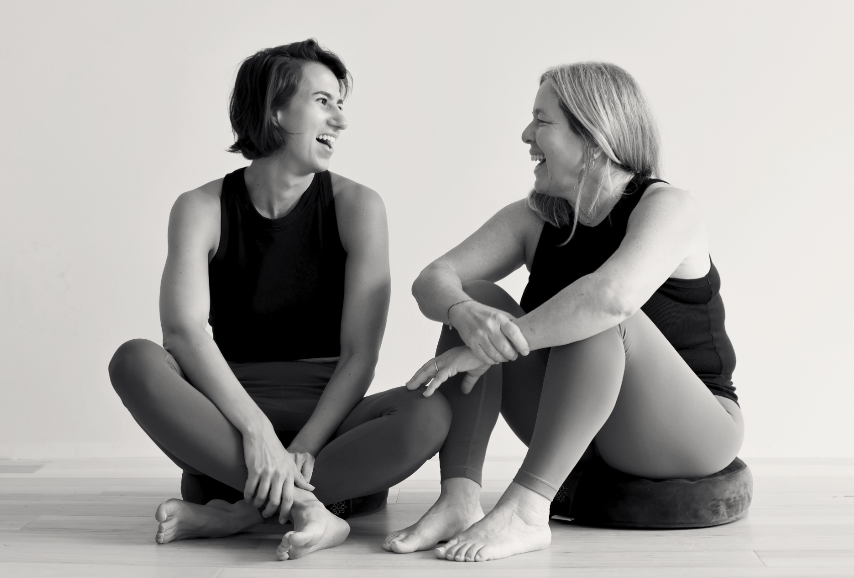 Two women sitting on the floor of a yoga studio, smiling and laughing.