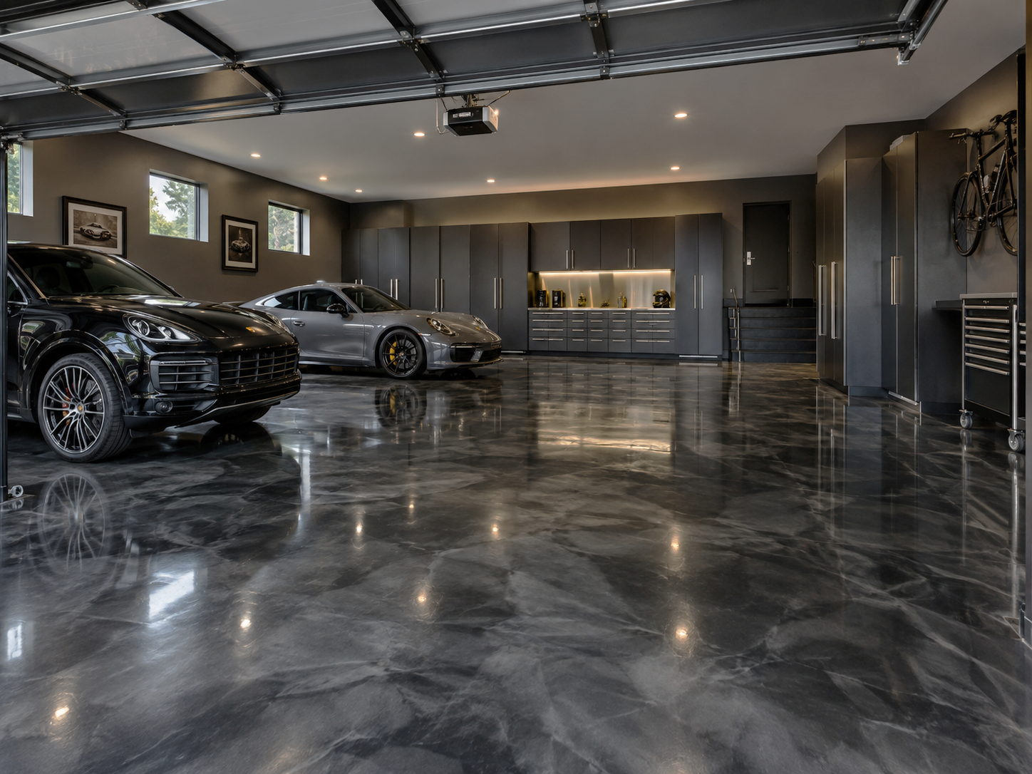 A modern, spacious garage with polished dark marble floors, three luxury cars including a black SUV, a silver sports car, and a gray sports car, gray built-in storage cabinets, wall artwork, windows, and a wall bicycle.