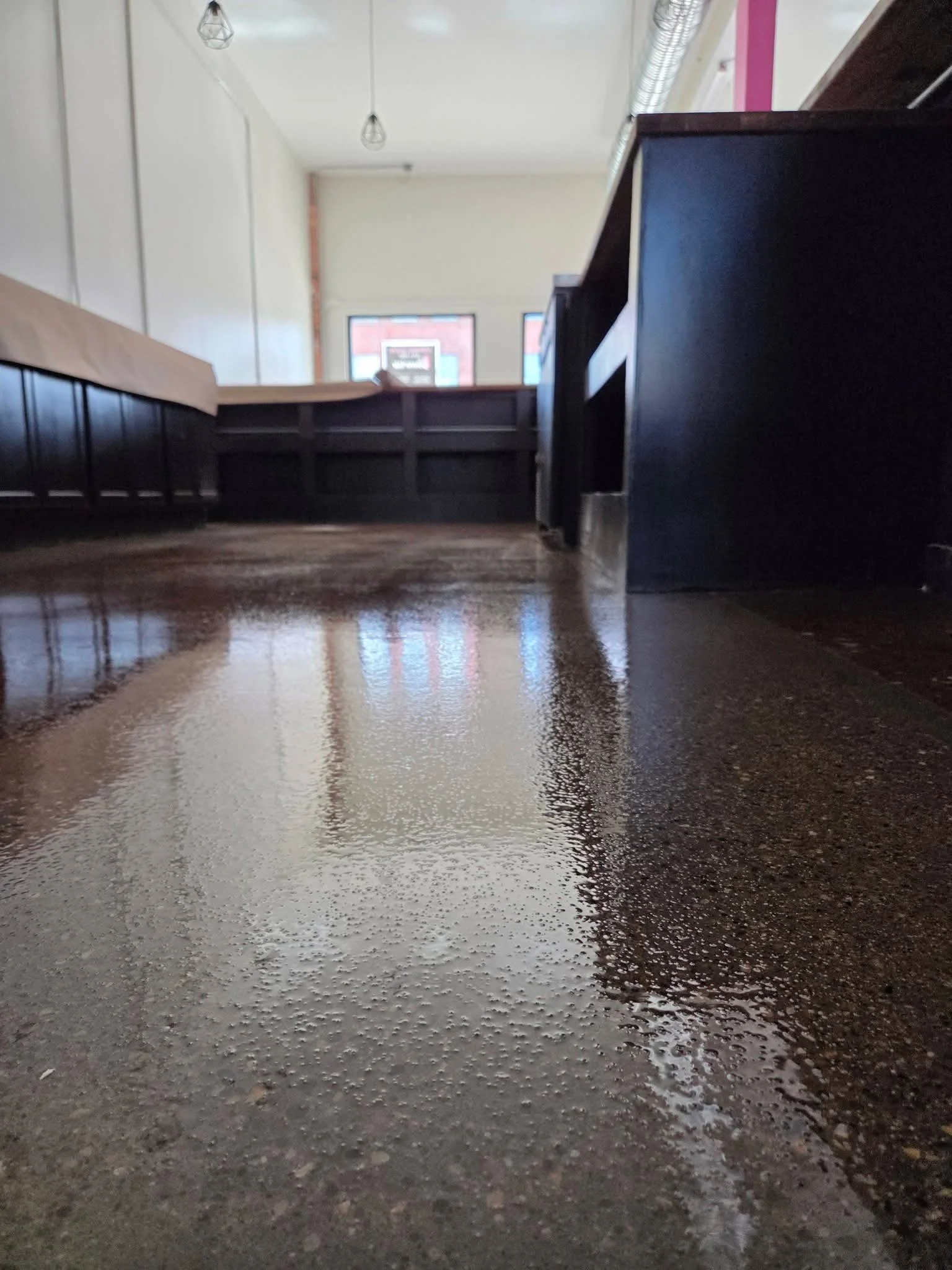 Interior of a building with a reflective, wet, brown floor, black furniture on the right, a white wall on the left, and windows in the background with a sign visible outside.