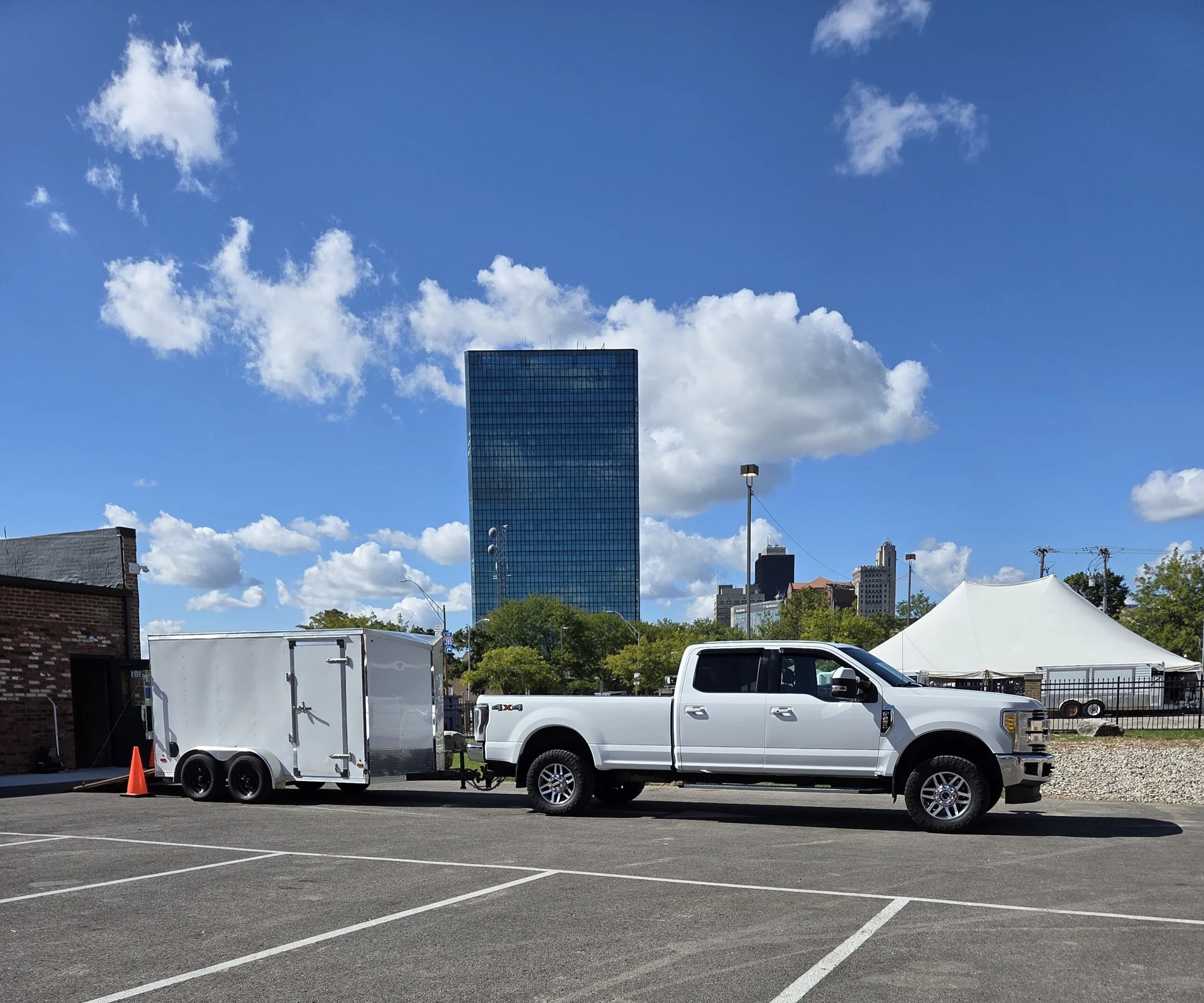 A white pickup truck with a trailer hitched to it, parked in an empty parking lot under a blue sky with scattered clouds. There is a white tent and some orange traffic cones nearby, with a city skyline in the background.