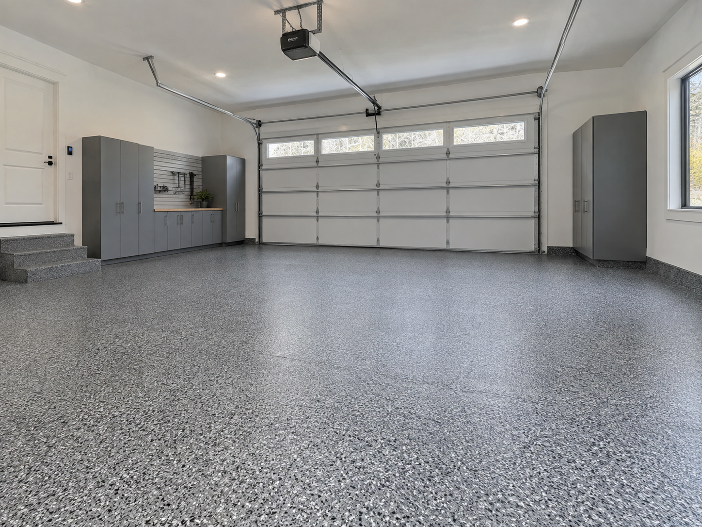 Empty clean garage with speckled gray epoxy floors, gray storage cabinets, a window, and a closed sectional garage door.