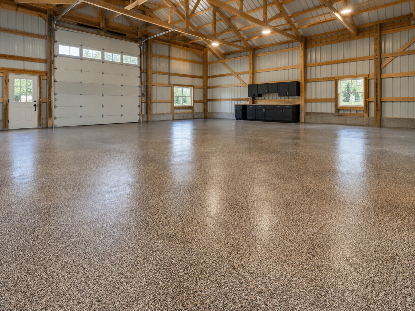 Empty garage with polished, speckled concrete floors, wooden beams, and metal walls. Features a large white sectional garage door, a smaller entry door, two windows, and black cabinets mounted on the wall.
