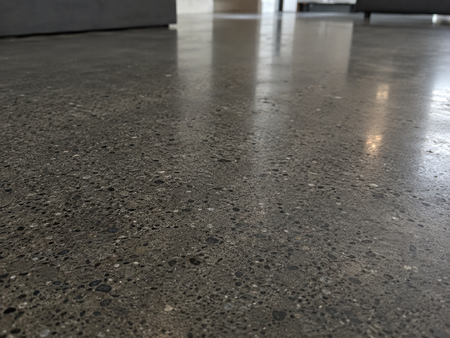 Close-up of a polished, concrete floor with a reflective surface, in an indoor setting with furniture visible in the background.