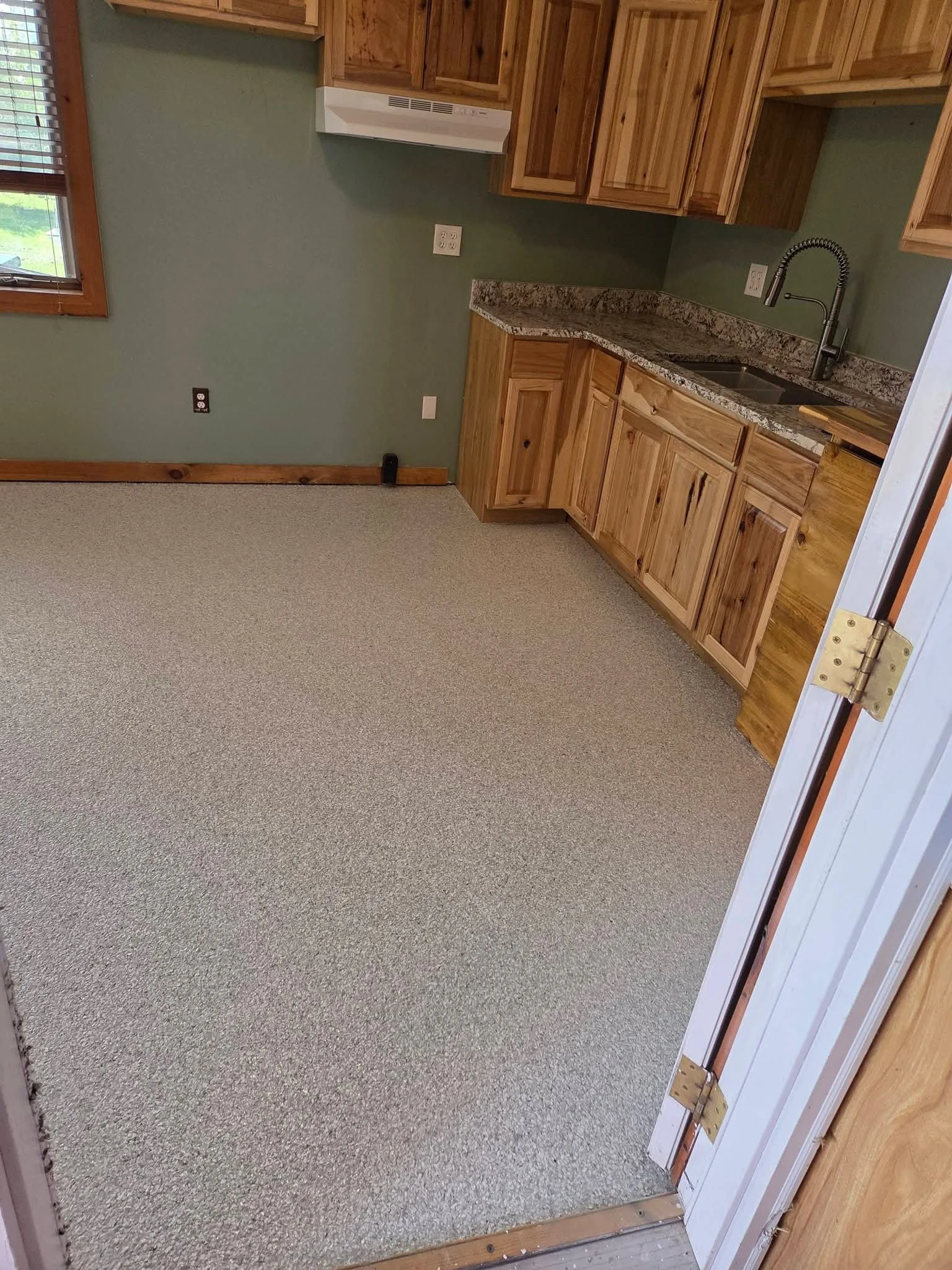 Kitchen with wooden cabinets, granite countertops, sink with modern faucet, and a window with blinds, with an open doorway leading to a room with beige carpet.