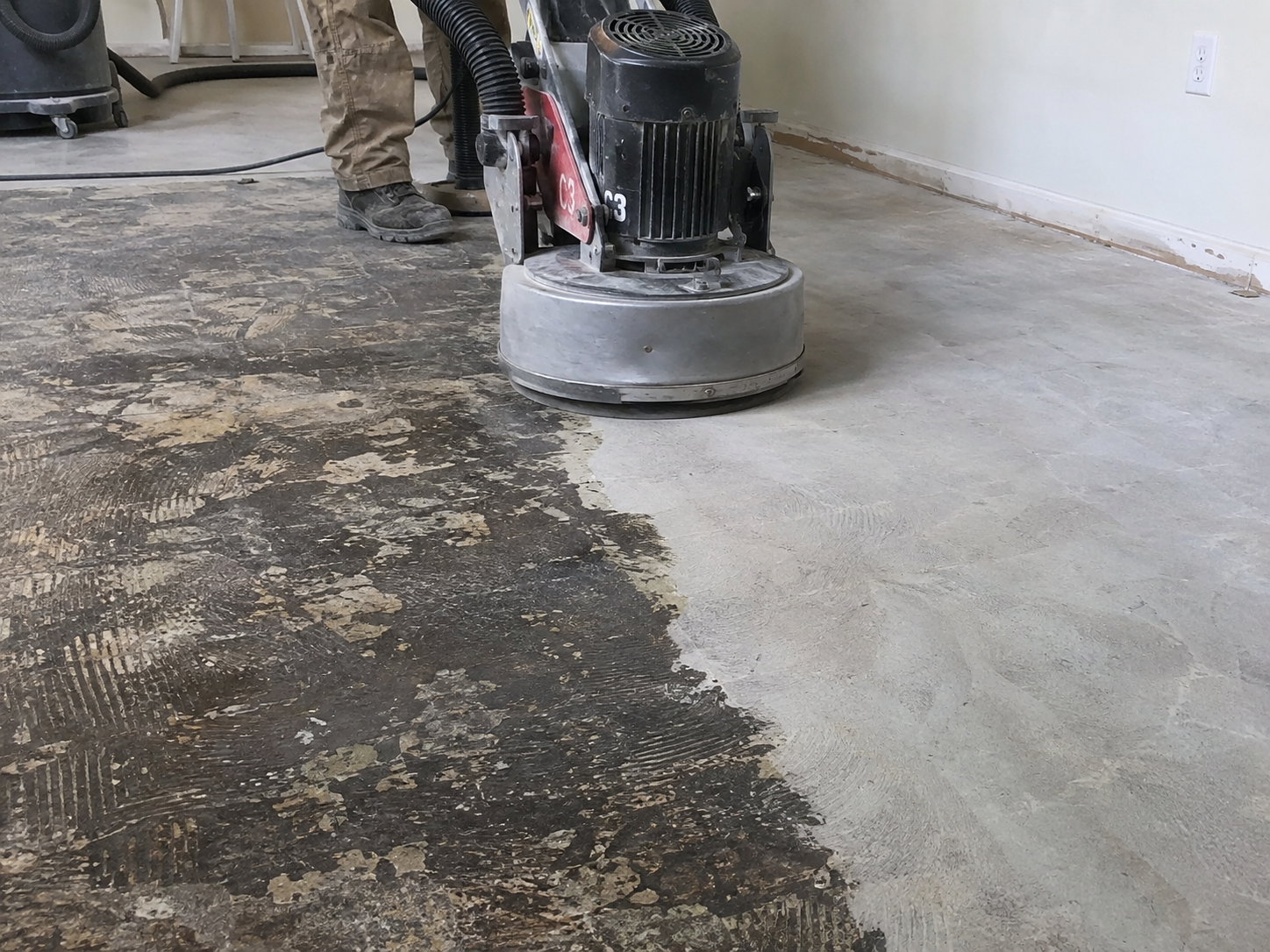 A worker is using a floor grinder on a concrete floor, grinding a section to remove old coating or prepare for a new finish. The floor is partially ground, showing a contrast between a darker, rougher area and a lighter, smoother area.