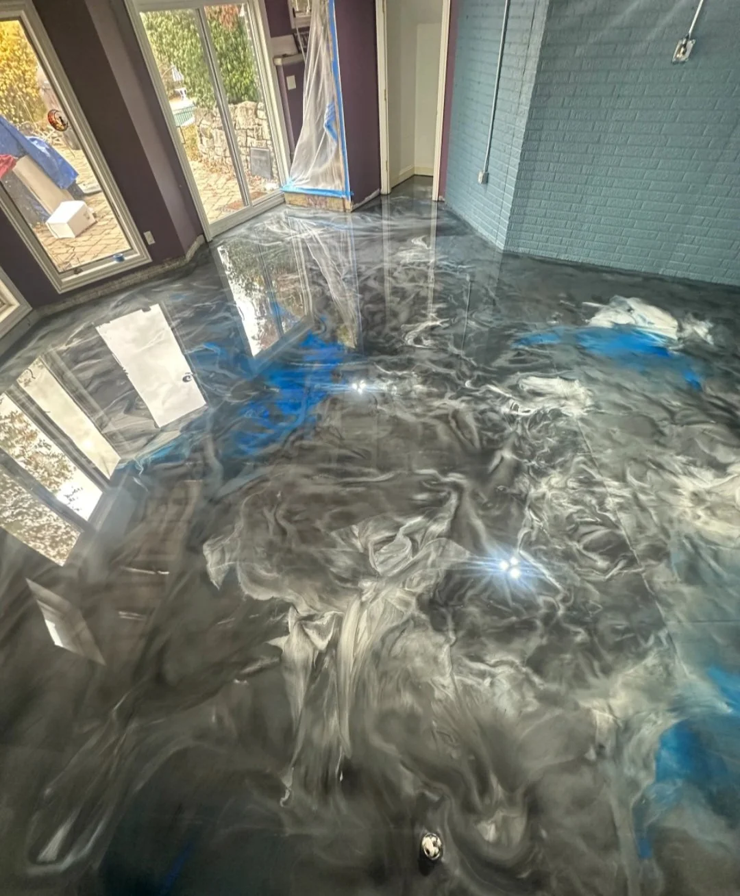 Interior space with a glossy, swirling black, white, and blue epoxy floor, near large window and sliding glass door, with a brick accent wall.