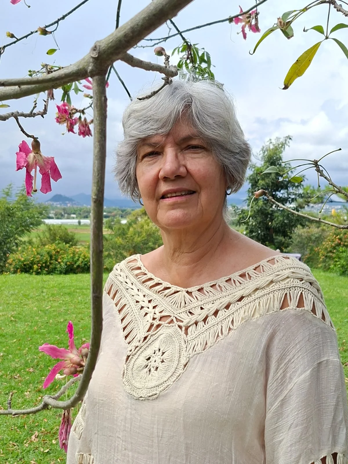 Anciana de cabello gris y blusa beige con bordados, de fondo un paisaje rural con árboles y montañas, en un día nublado.
