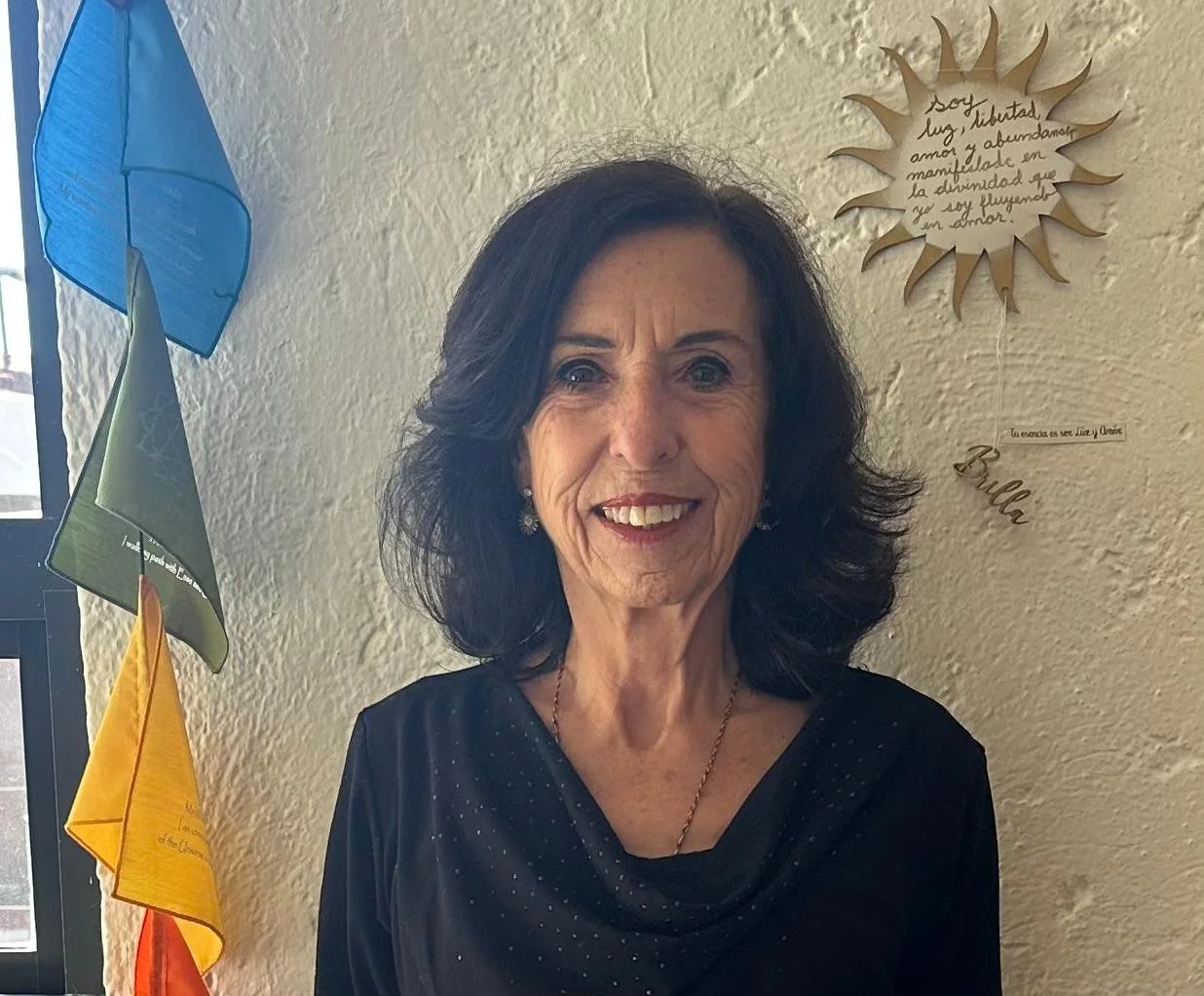 Retrato de una mujer con cabello oscuro y rizado, viste una blusa negra y sonrie frente a una pared con decoración de papel y banderas coloridas, en un lugar con ventanas que dejan entrar luz natural.