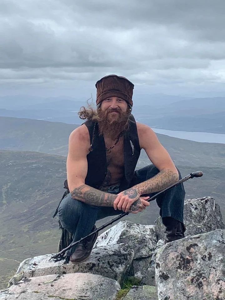 Un hombre con barba, tatuajes y gorro de lana posa en la cima de una montaña, sosteniendo un bastón, con un paisaje montañoso y un lago en el fondo bajo un cielo nublado.
