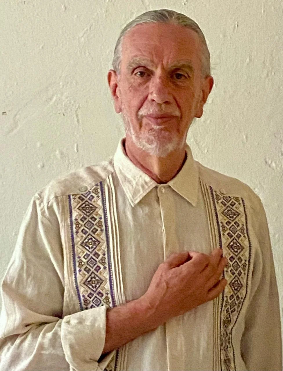 Hombre mayor con cabello y barba gris, vistiendo una camisa blanca con bordados tradicionales en los lados, sosteniendo su mano en el corazón delante de una pared de color claro.