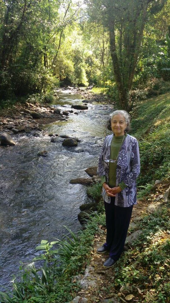 Una mujer mayor de cabello rizado gris, vestida con una blusa verde y una chaqueta de diseño, de pie junto a un río rodeado de árboles y vegetación en un entorno natural.