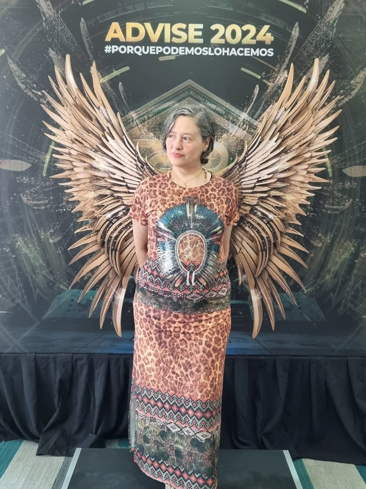 Mujer con cabello rizado y corto, vestida con una blusa de estilo indígena y falda larga, de pie frente a un fondo decorativo con alas y un cartel que dice 'ADVISE 2024 #PORQUEPODEMOSLOHACEMOS'.