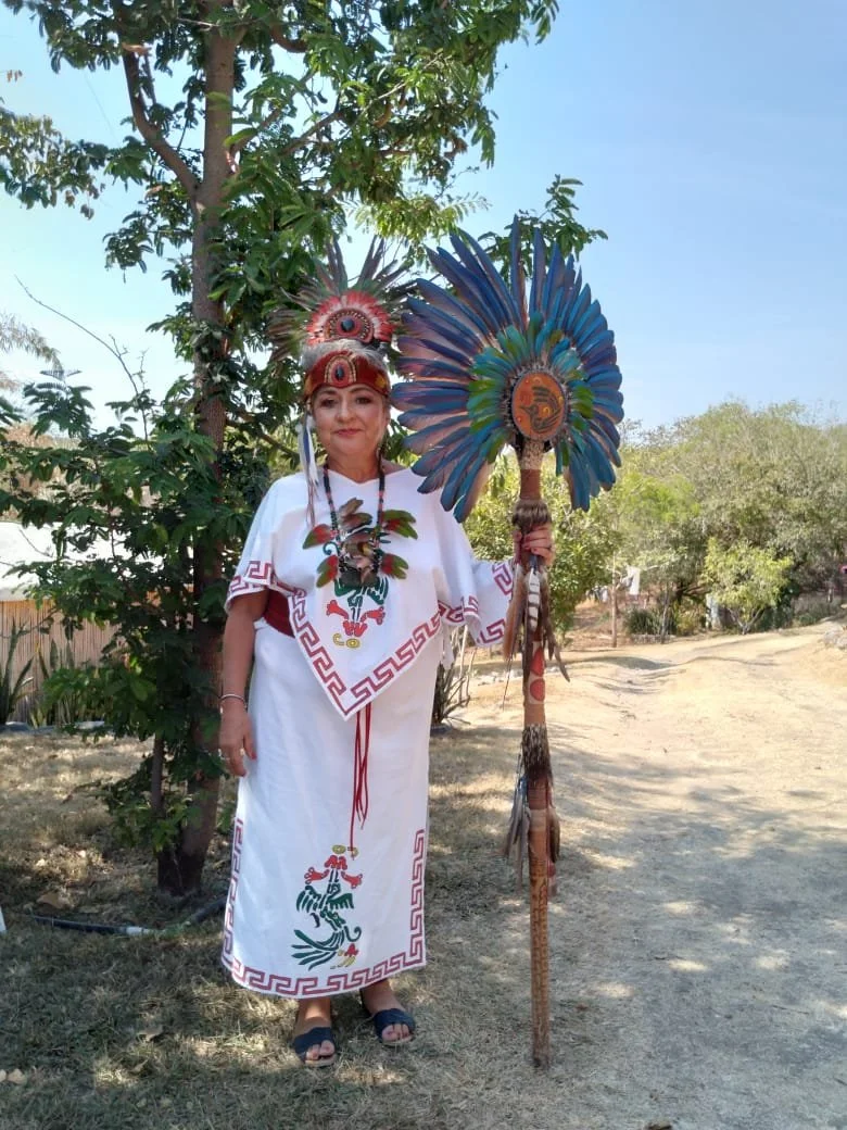 Mujer vestida con ropa tradicional, usando un tocado y sosteniendo un tambor ceremonial con plumas en un entorno al aire libre.