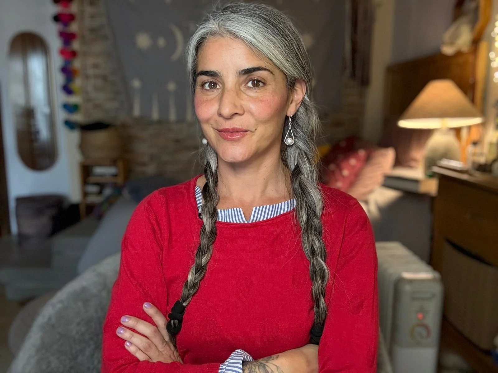 Retrato de una mujer con cabello gris y trenzado, usando una camiseta de rayas debajo de un suéter rojo, en un ambiente hogareño con lámpara y muebles en el fondo.