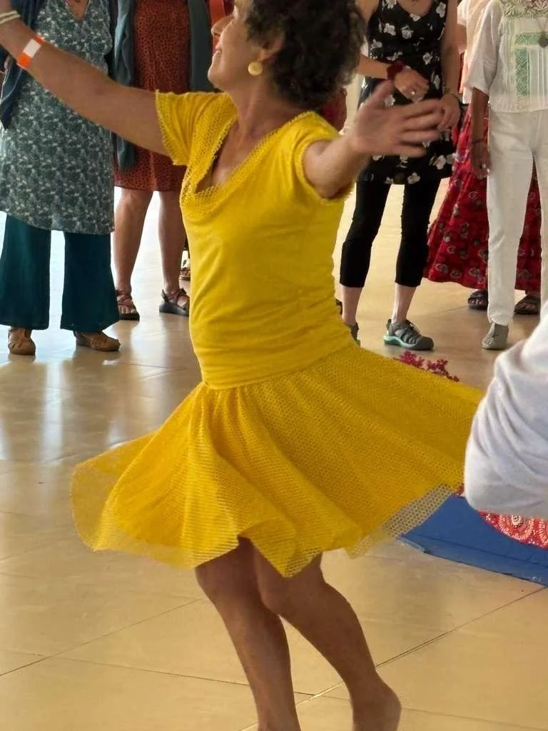 Mujer bailando con vestido amarillo, rodeada de otras personas en un espacio interior.