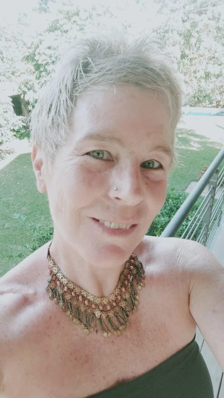 Mujer con cabello corto rubio, usando collar decorativo, en un balcón con vista a un jardín verde y una piscina en el fondo.