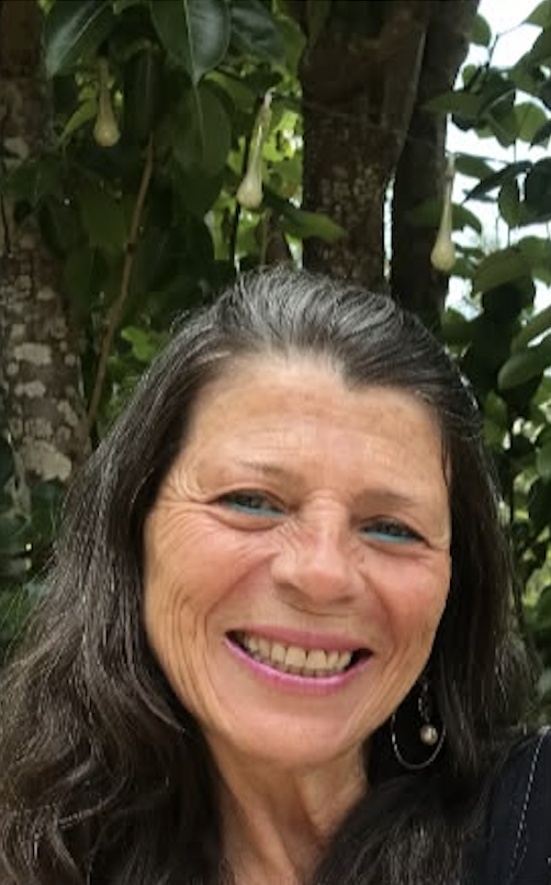 Mujer sonriendo con cabello largo oscuro, en un entorno natural con árboles y flores blancas colgantes.
