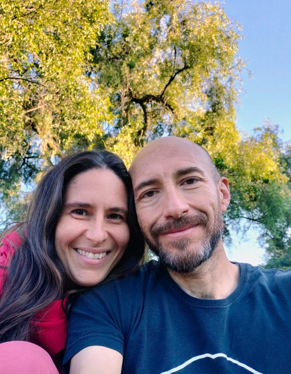 Una pareja sonriendo, tomando una selfie al aire libre con árboles verdes y cielo azul en el fondo.