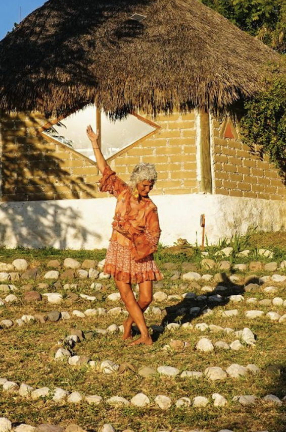 Una mujer mayor con cabello gris, vestida con ropa de color naranja y una falda de estampado en tonos similares, bailando en un espacio al aire libre con rocas en el suelo, frente a una casa con paredes de ladrillo y un techo de paja.