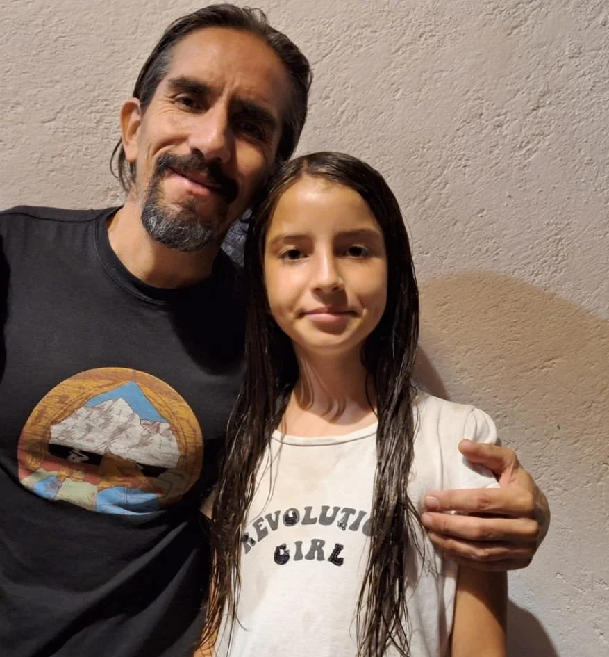 Un hombre y una niña posando juntos frente a una pared de color beige, el hombre lleva una camiseta negra con un diseño de una montaña, la niña lleva una camiseta blanca que dice "Revolution Girl".