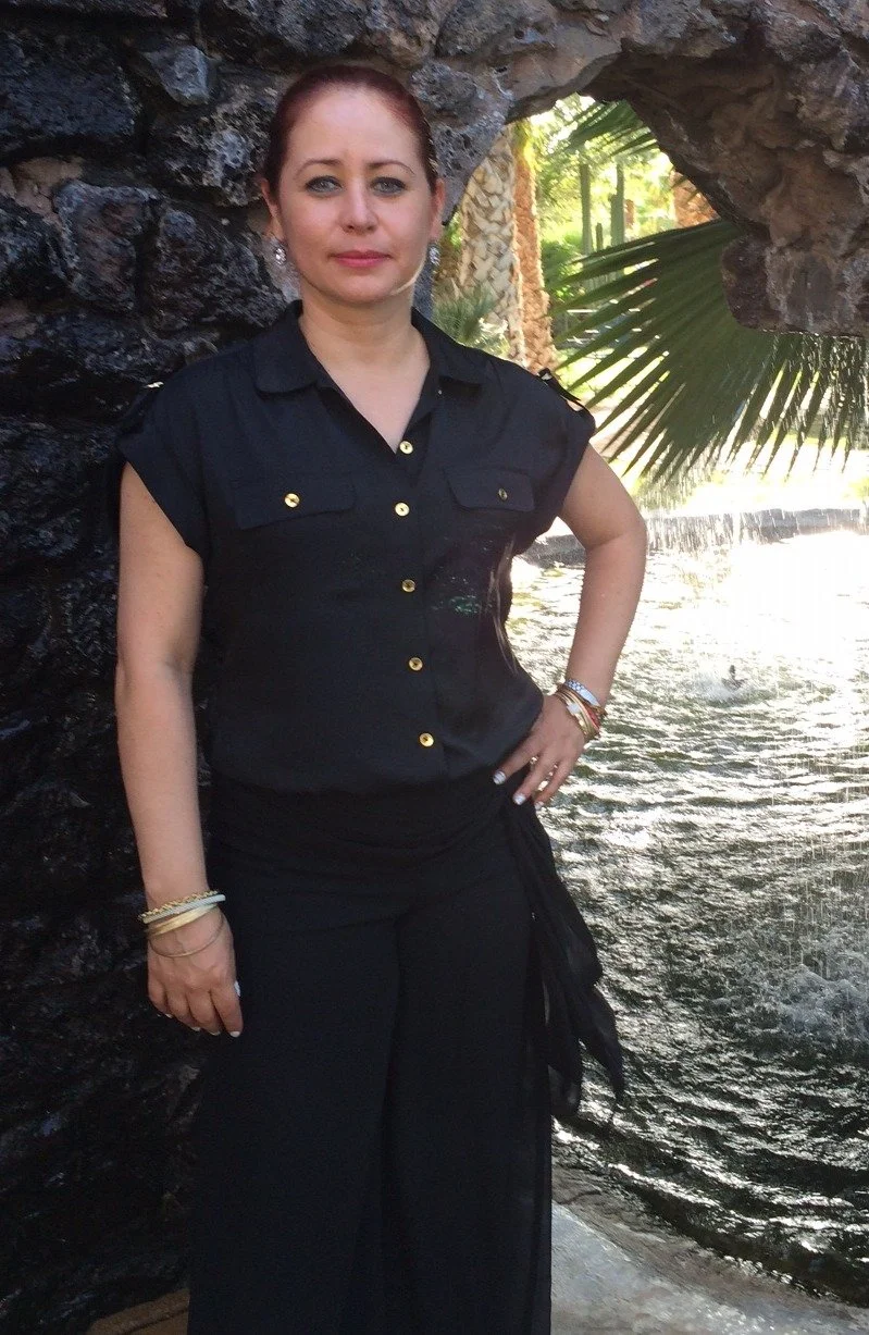 Mujer con cabello castaño claro y ropa negra, posando junto a una roca en un entorno de naturaleza con palmeras y agua en el fondo.
