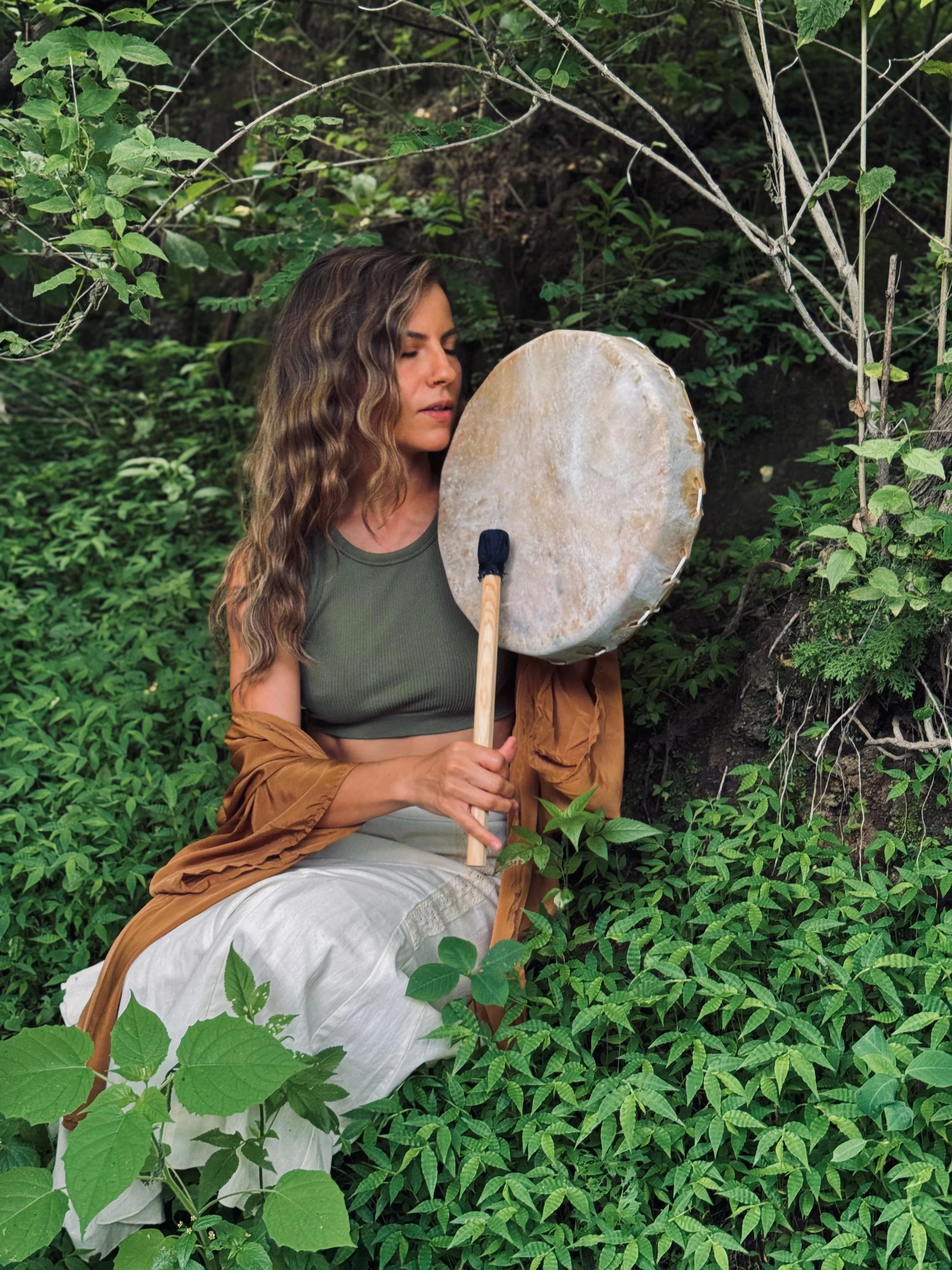 Mujer con cabello rizado sosteniendo un tambor en medio de vegetación verde densa en un entorno natural.