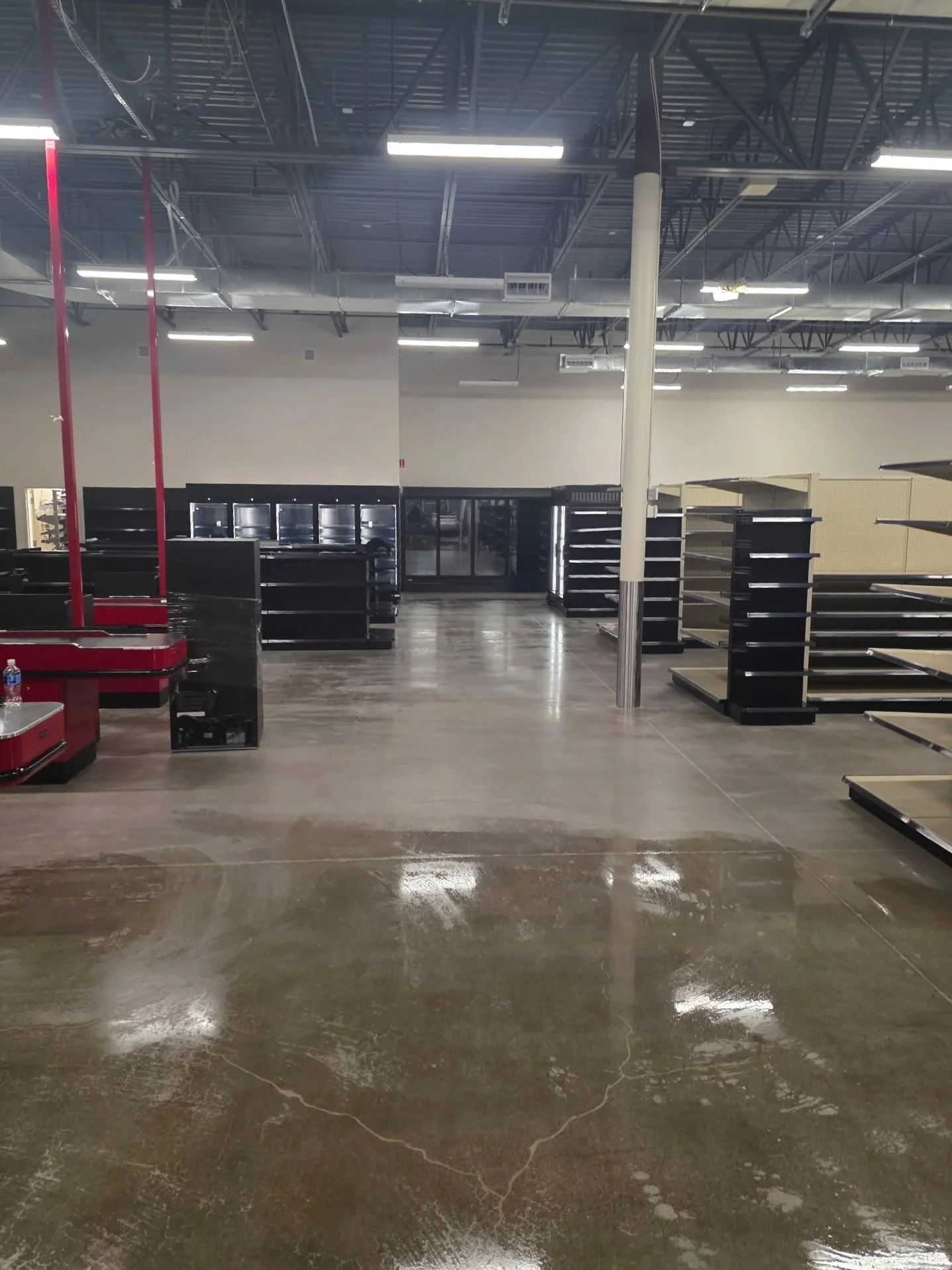 Empty retail store with black metal shelving units, some with glass doors, and a red checkout counter, with a polished concrete floor and visible ceiling piping and lighting fixtures.