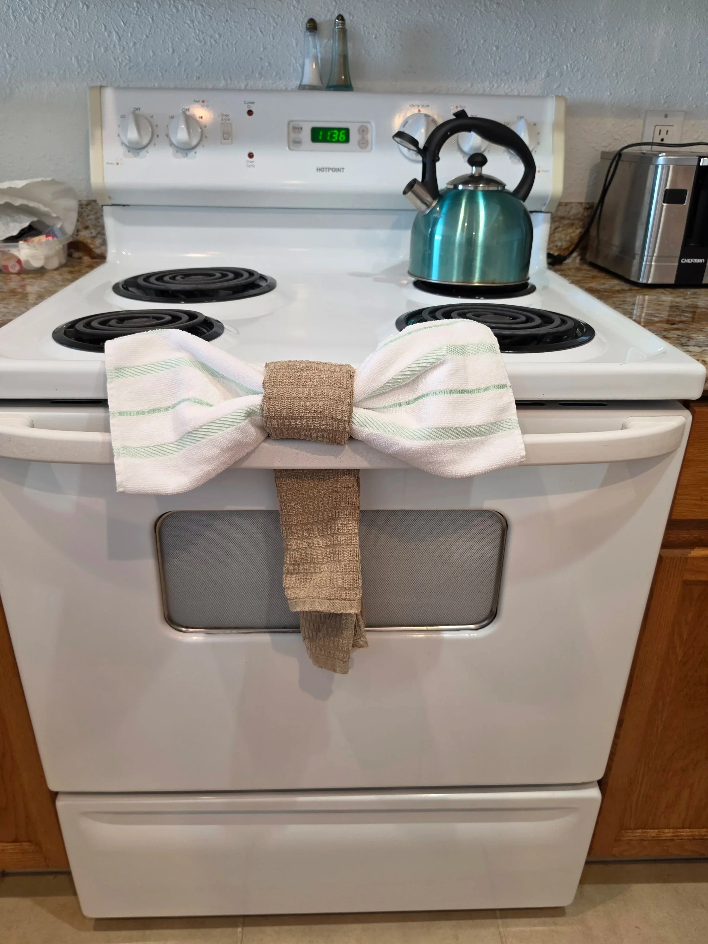 White electric stove with four burners, a teal kettle on the right front burner, a white and green striped cloth with a brown towel on top hanging from the oven handle, a granite countertop, and a stainless-steel toaster on the right side.