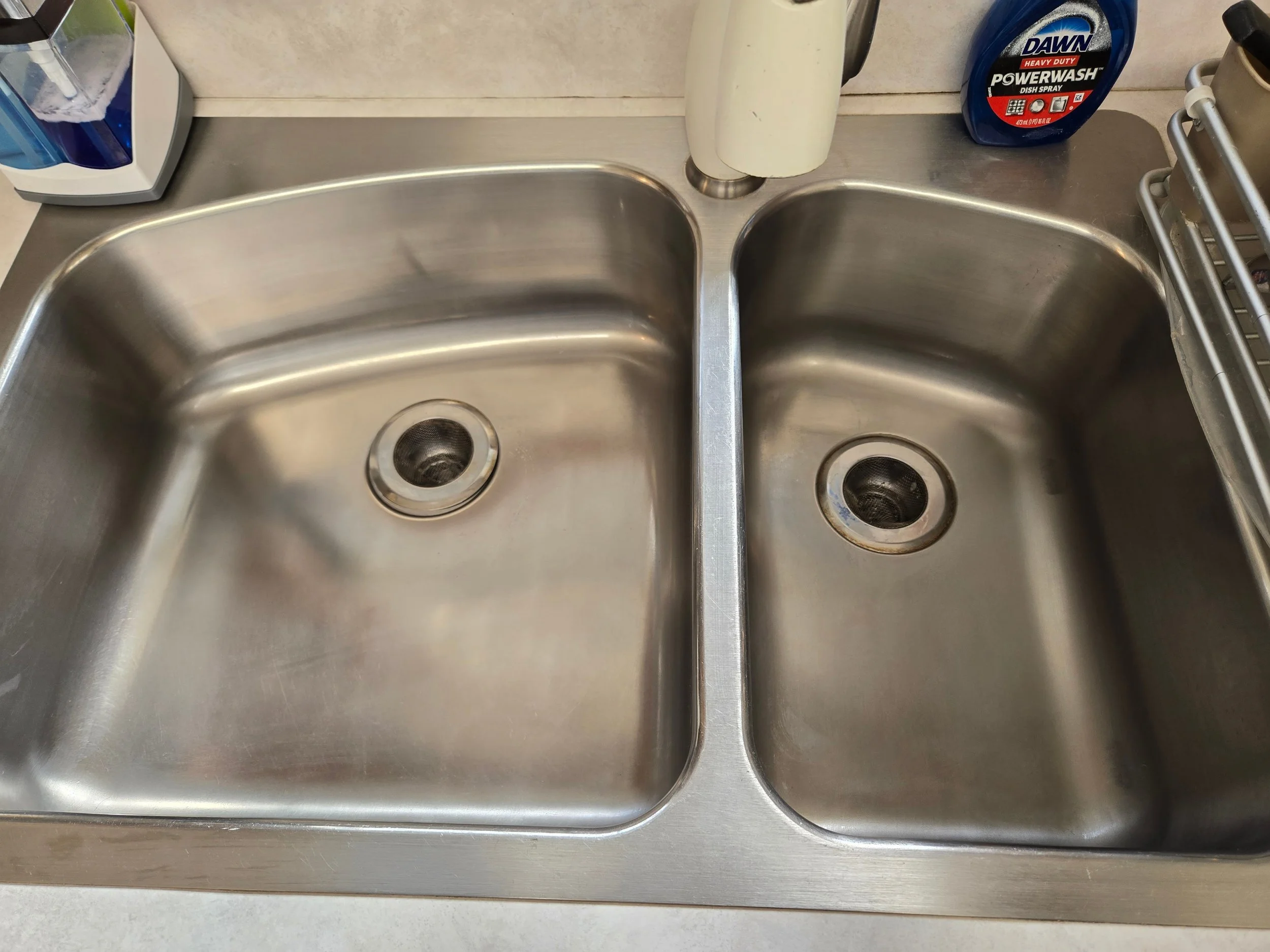 Stainless steel double kitchen sink with a sticker of Dawn dish soap on the right side and a paper towel holder in the middle, with a partially visible dish soap bottle on the left.