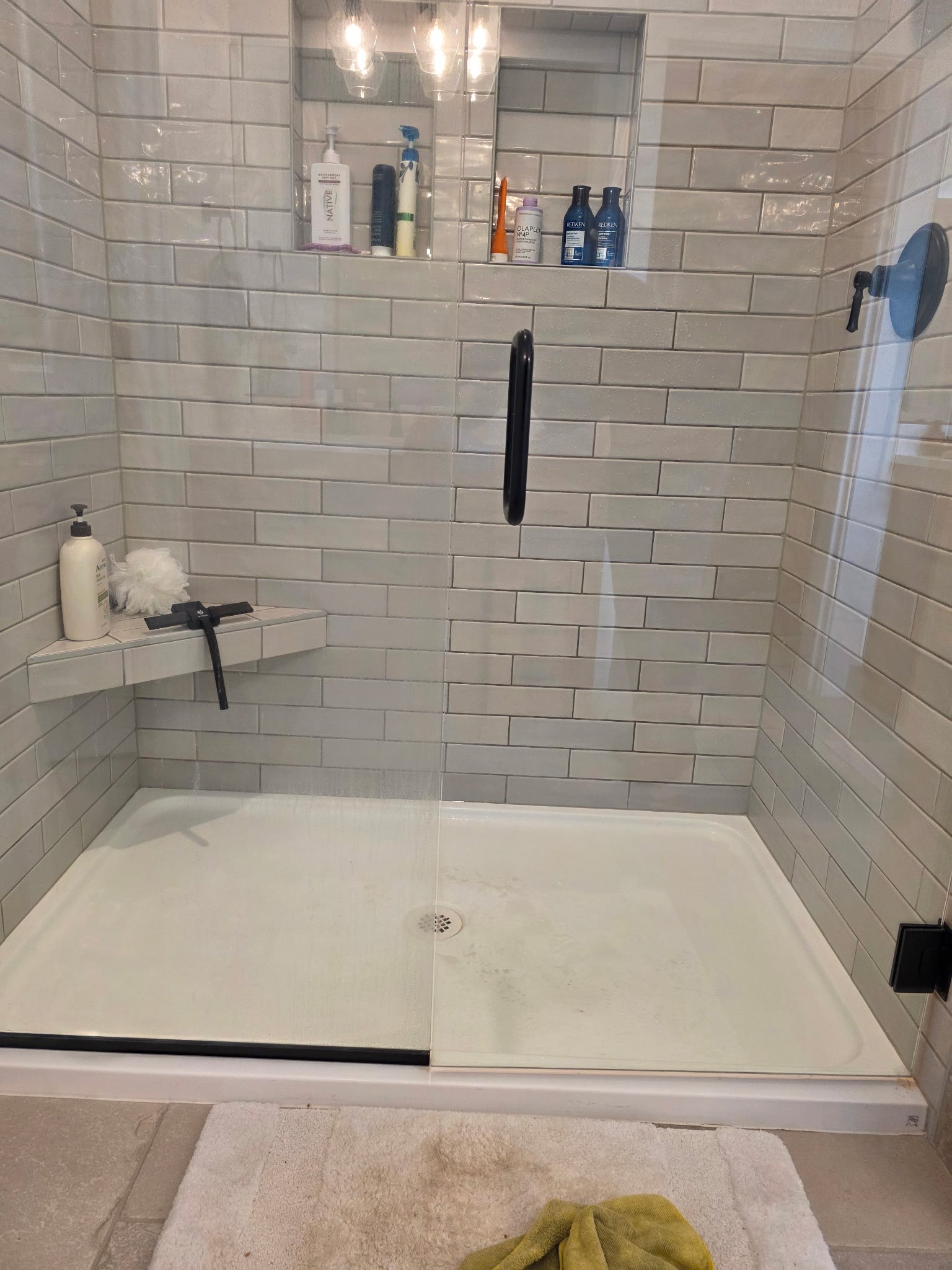 Shower with beige tiled walls, a built-in shelf with toiletries, a glass door, and a white bath mat on the floor. A green towel is on the floor near the mat.