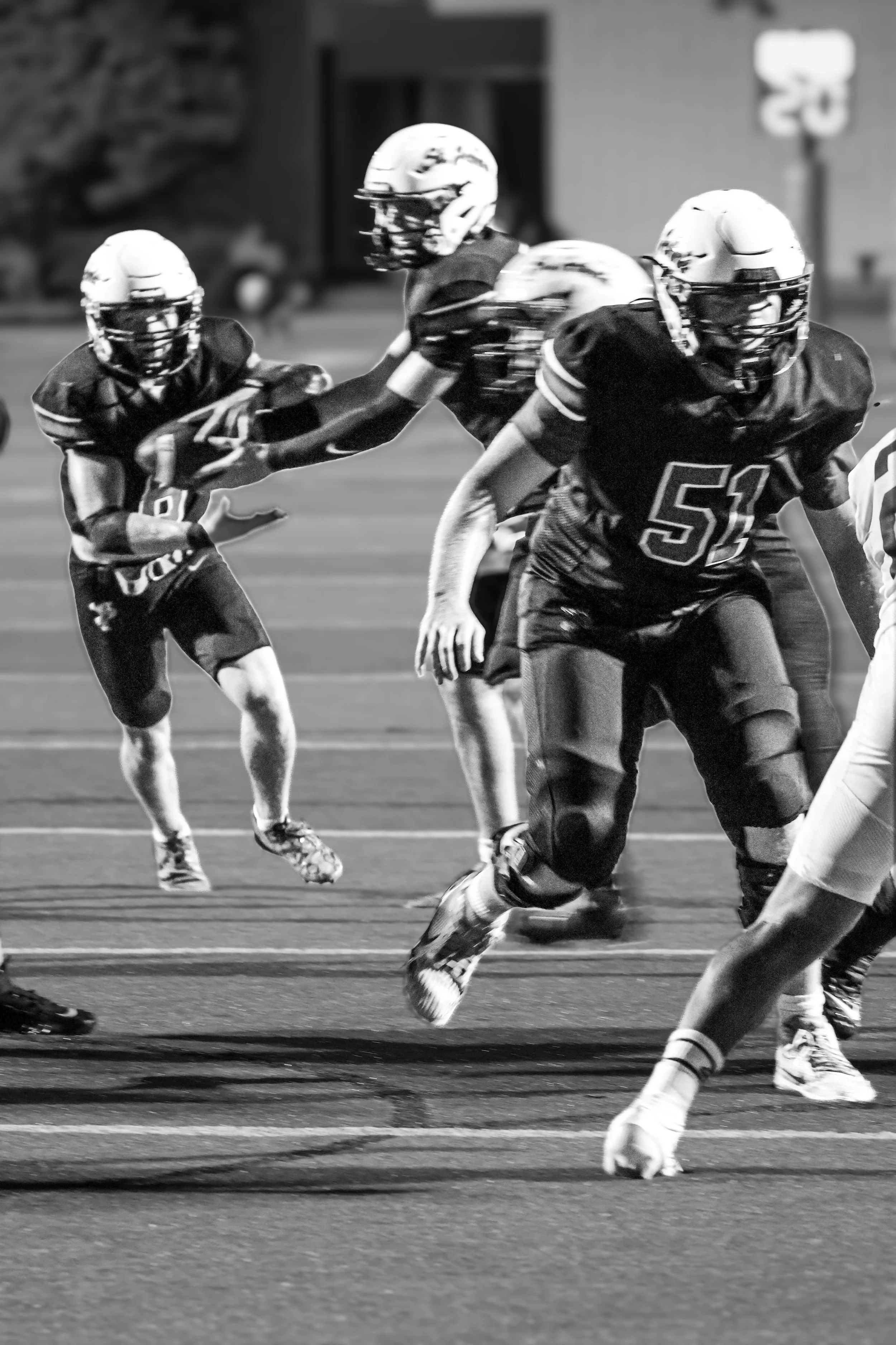 Black and white action shot of an American football game showing players running on the field, with some reaching for the ball and others protecting it.