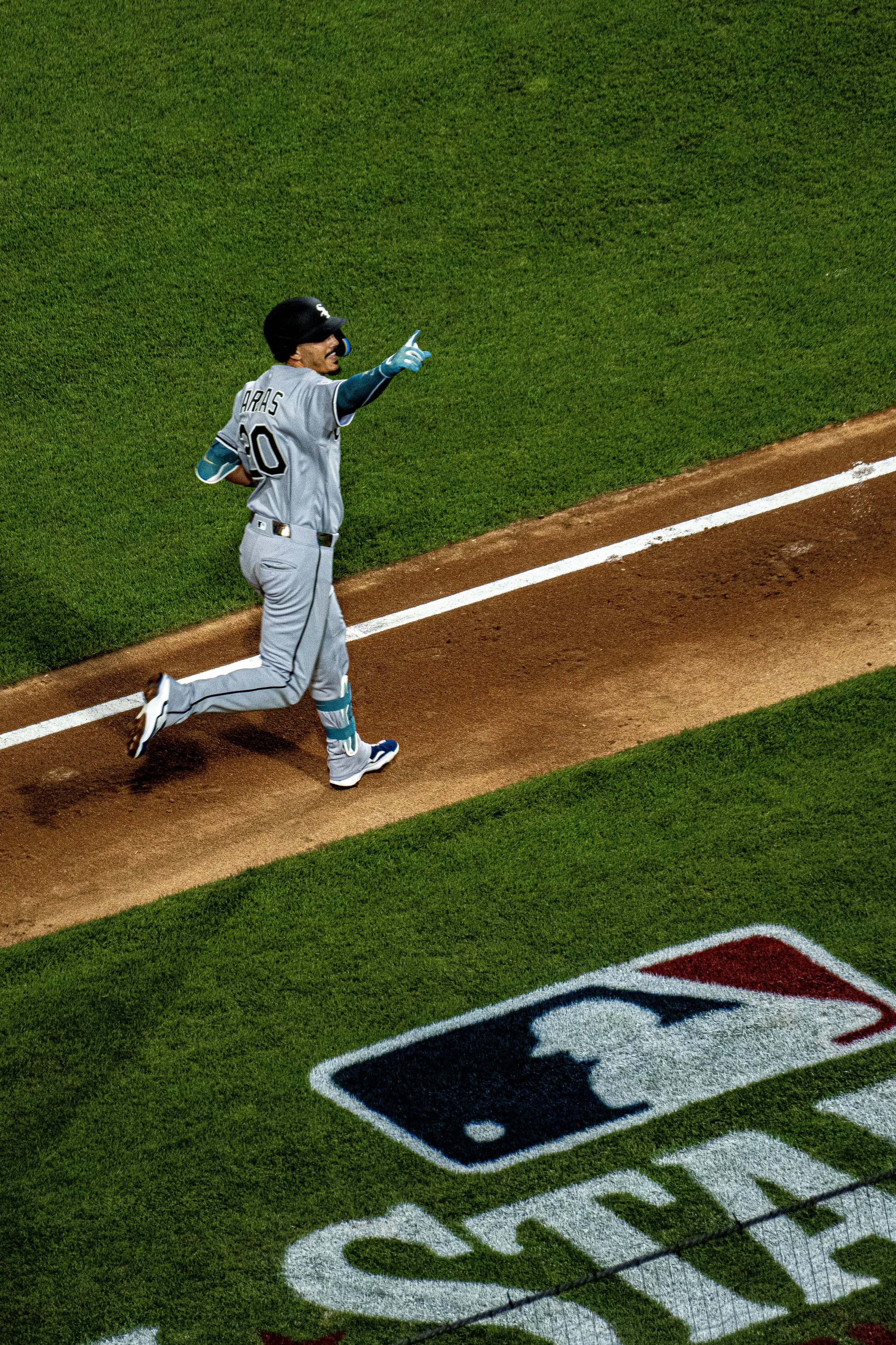 A baseball player in a gray uniform with the number 20, wearing a black helmet and blue gloves, celebrating on the field near the scoreboard with the Major League Baseball logo visible on the ground.